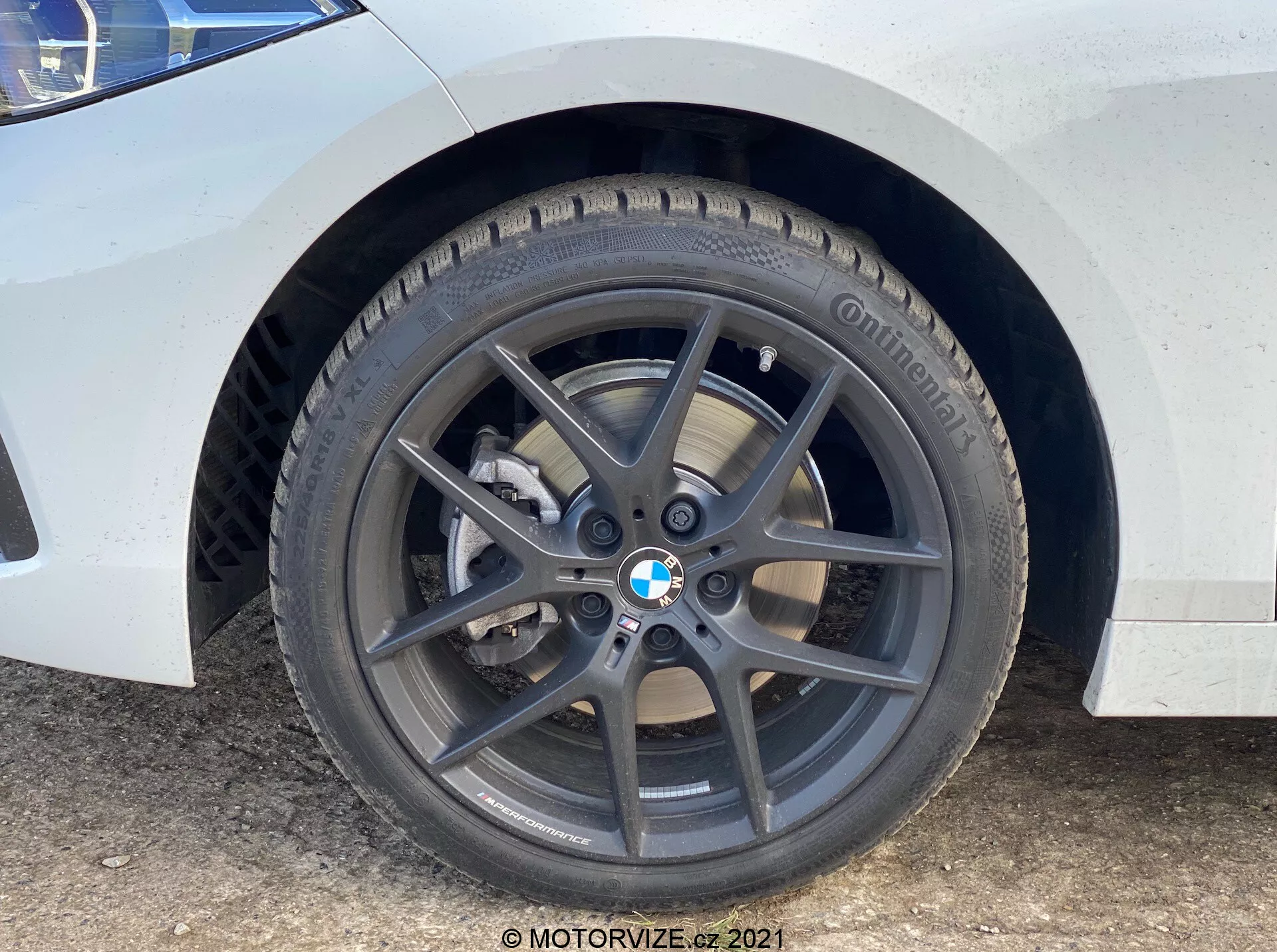 Gros plan sur la roue avant et l'aile du BMW Série 1 2019 (BMW-116i). La perspective au niveau du sol met en valeur la roue en alliage à cinq branches gris foncé avec l'emblème BMW, le pneu Continental avec bande de roulement visible, le disque de frein ventilé et l'étrier de frein à piston unique argenté. La carrosserie blanche avec des taches de saleté et d'eau indique une utilisation active.