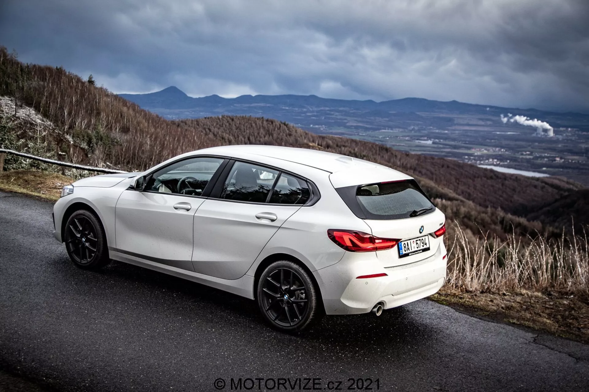 Vue latérale arrière en angle d'une BMW 116i hatchback blanche de 2019 sur une route en asphalte avec un ciel dramatique couvert. L'image met en avant le design arrière du véhicule, les arches de roues arrière, les jantes en alliage sombres, le système d'échappement double et le profil latéral incluant les portes passager arrière et conducteur. Le décor suggère une route collineuse ou montagneuse, mettant en avant les attributs sportifs et dynamiques de la voiture. Présence du filigrane du photographe 'MOTORWIZE.cz 2021'.