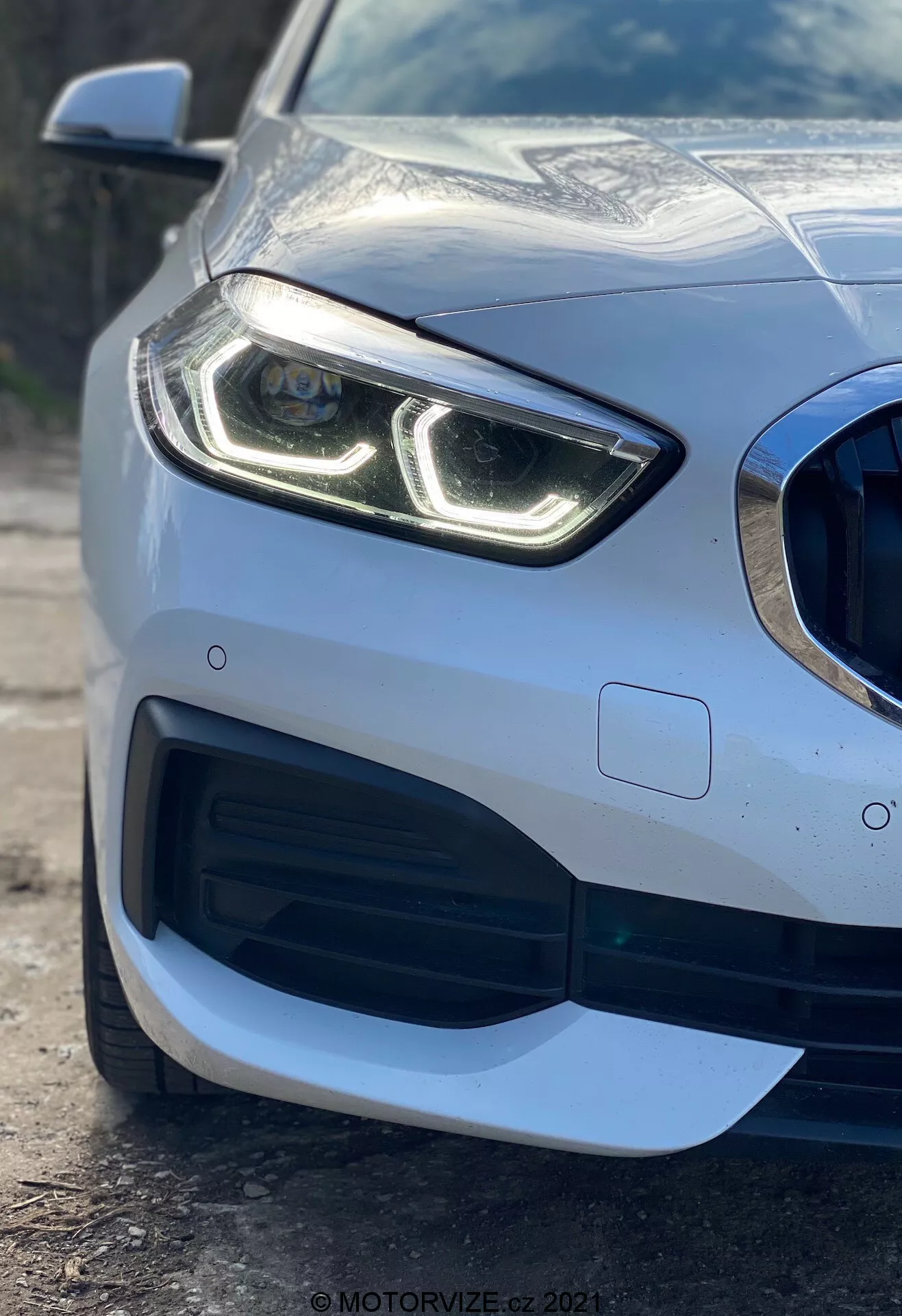 Vue rapprochée de la section avant gauche d'une voiture BMW Série 1 2019 (BMW 116i) mettant en avant son phare moderne avec feux de jour à LED distinctifs, grille en nid d'abeille avec grande prise d'air, design aérodynamique sophistiqué du pare-chocs avant et détecteur de parking intégré dans le pare-chocs, montrant la finition de peinture métallique premium et la carrosserie réfléchissante.