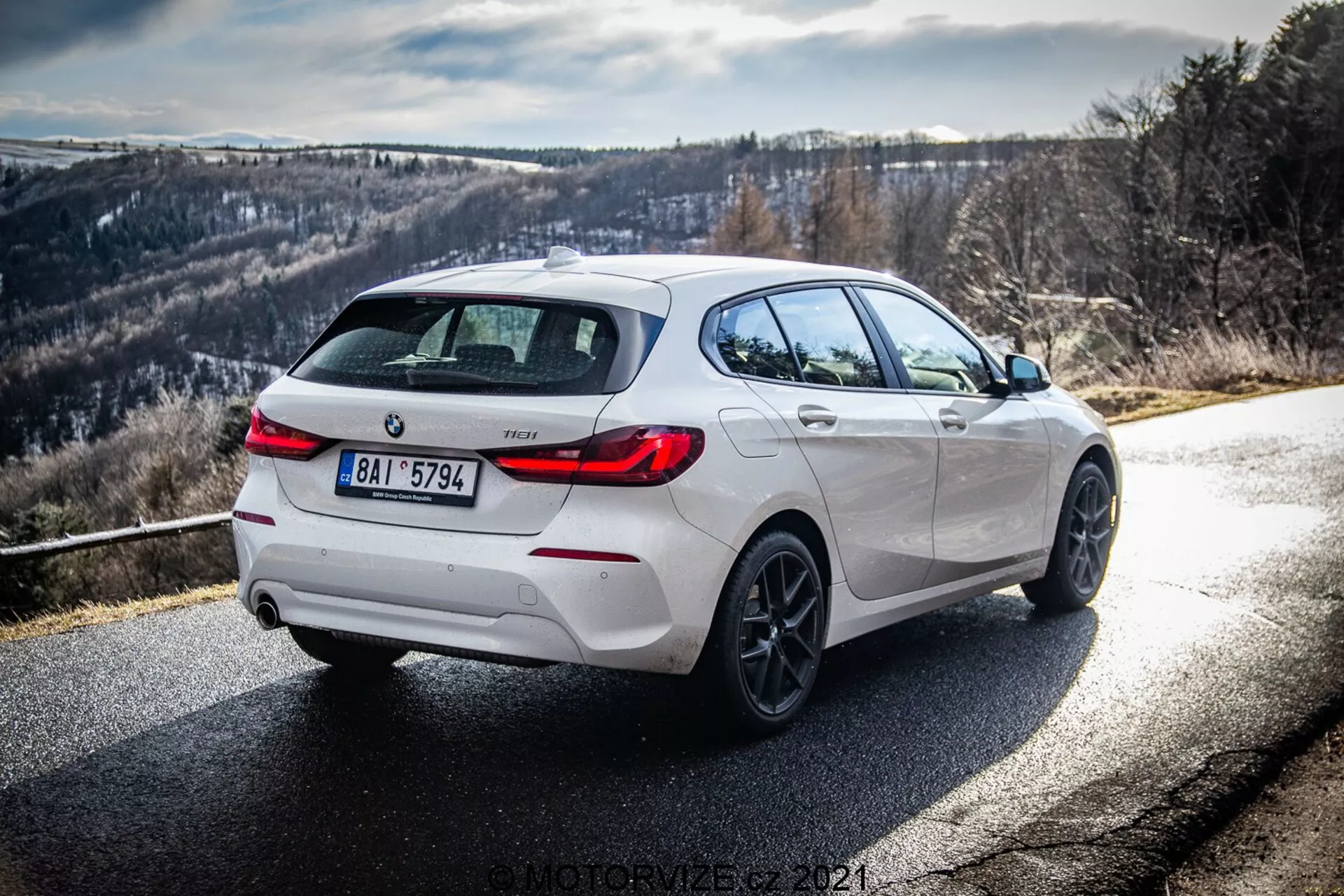 Vue arrière trois-quarts dynamique d'une BMW 116i 2019 blanche, montrant son côté droit et arrière sous la lumière du jour avec de clairs rayons de soleil et des ombres. L'image souligne les détails de conception, y compris les accents noirs sur le diffuseur arrière, les jantes et les coques de rétroviseurs latéraux, complétés par des roues en alliage et un système d'échappement double, suggérant une variante plus sportive. La route vire à droite avec un arrière-plan de collines ou de montagnes, idéal pour les passionnés de conduite.