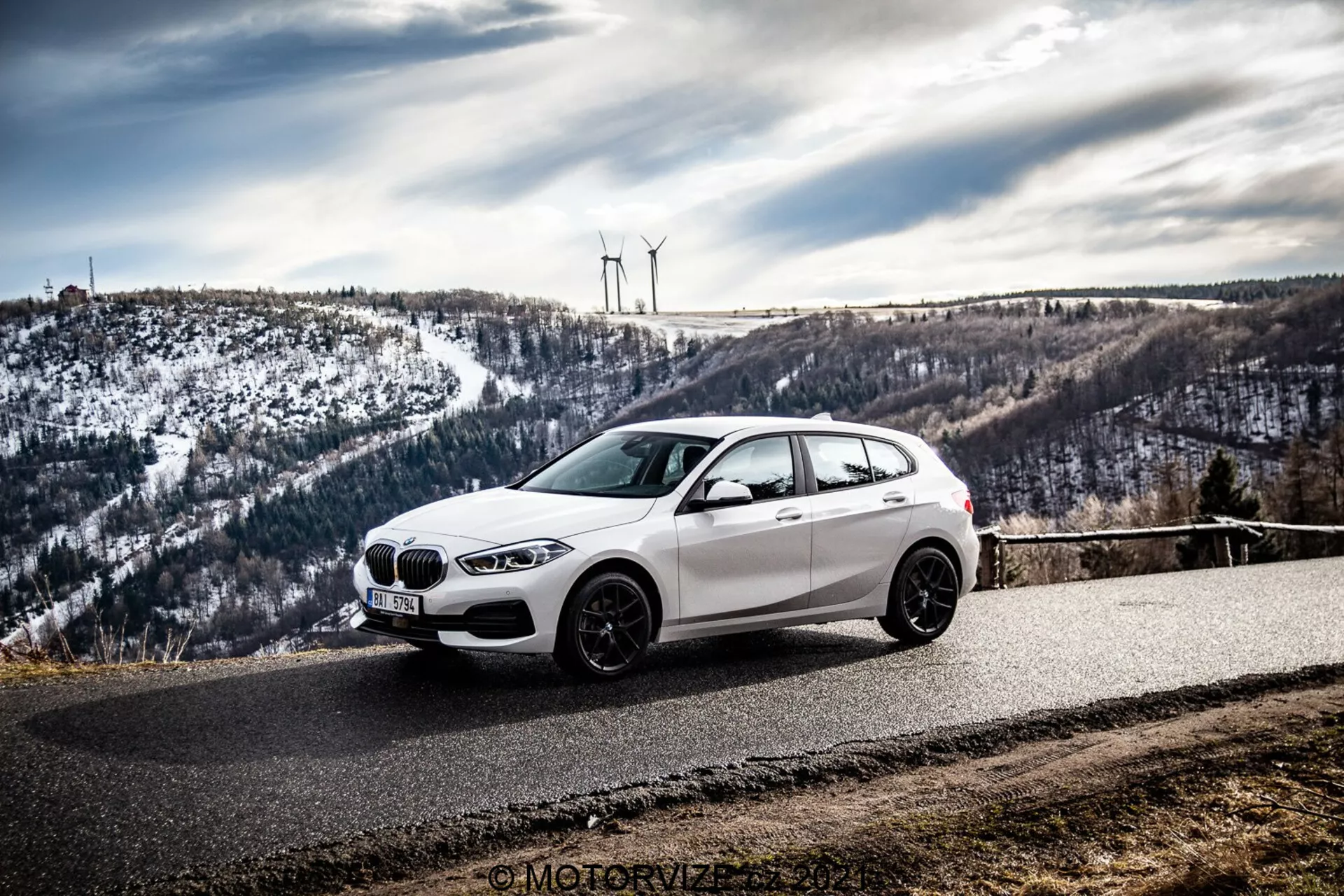 Une voiture à hayon BMW blanche, probablement une BMW 1er-Reihe 2019 (BMW 116i), est garée sur une route asphaltée en courbe entourée d'un paysage vallonné et enneigé avec des éoliennes. La photo, prise sous un angle avant-latéral, met en valeur la calandre avant, les phares, les lignes aérodynamiques et les roues en alliage de la voiture sous un ciel couvert.