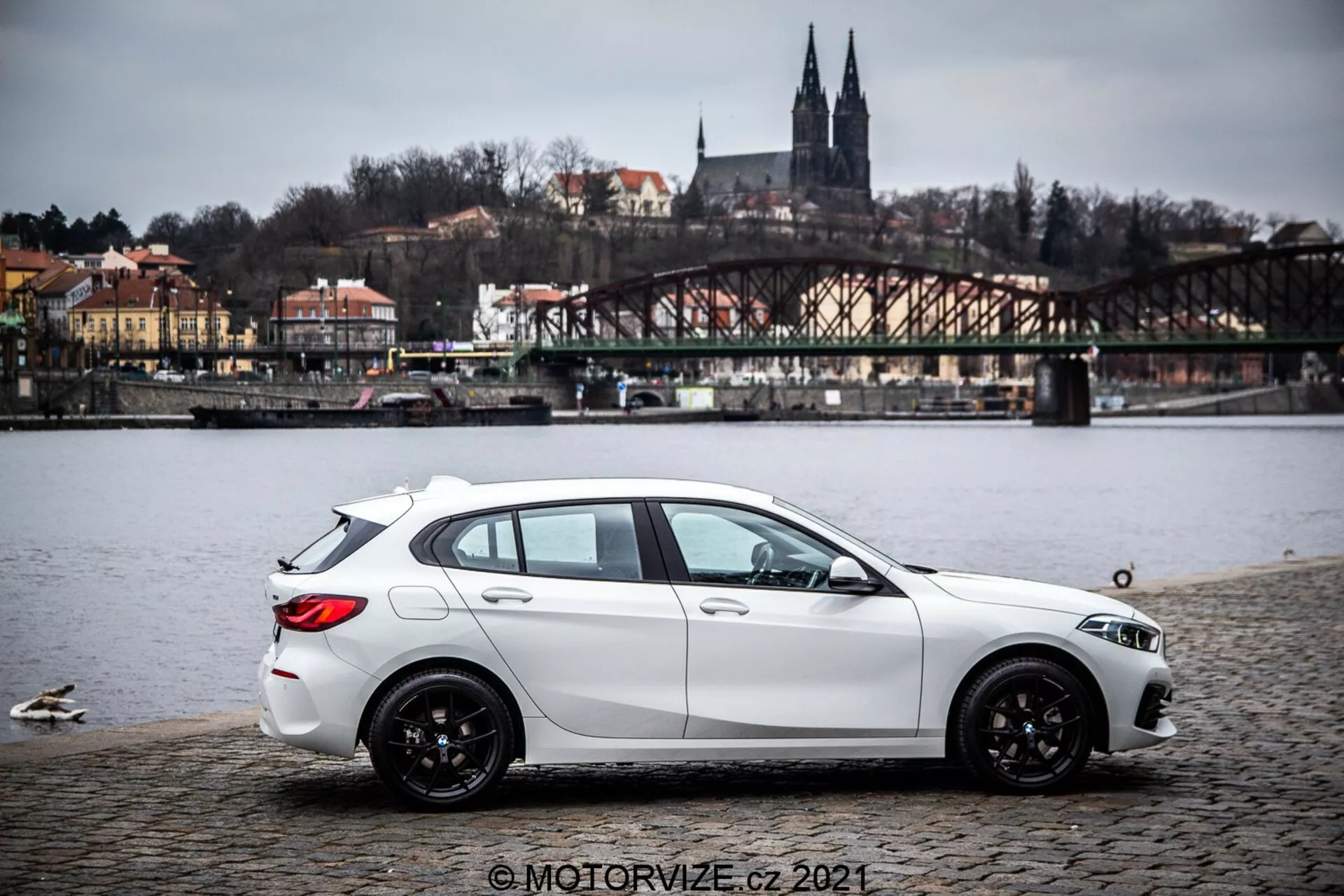 Une BMW Série 1 de 2019 (BMW-116i) blanche, garée sur une surface en pavés près d'un plan d'eau avec des canards, montrant un profil latéral avec une vue arrière légère. La voiture présente des lignes sculptées dynamiques, une ligne de toit inclinée, des roues en alliage noir à multiples rayons et des phares et rétroviseurs latéraux visibles. À l'arrière-plan un pont, des bâtiments sur une colline et des éléments architecturaux suggérant un cadre européen. L'éclairage est diffus sous un ciel couvert et l'image comprend un filigrane © MOTORVIZE.cz 2021.