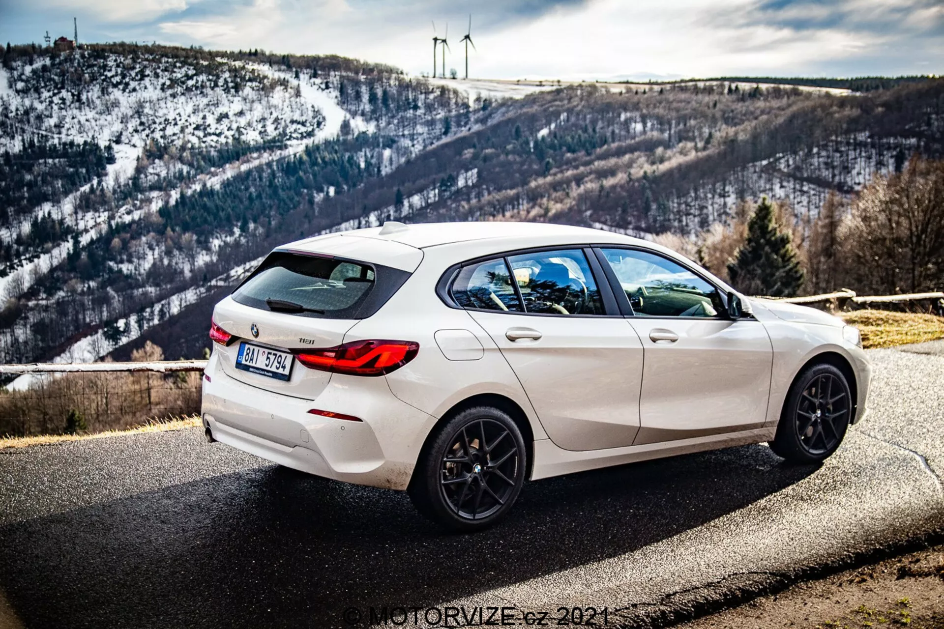 L'image montre une BMW Série 1 (BMW-116i) de 2019 blanche, garée en bord de route avec un paysage pittoresque en arrière-plan incluant un paysage vallonné couvert de neige et des éoliennes à l'horizon. La photographie est prise d'un angle de trois quarts arrière, exposant clairement le côté droit de la voiture. La vue montre les lignes de design élégantes, le spoiler arrière, les jantes noires à cinq branches, les doubles sorties d'échappement et les vitres teintées à l'arrière. La voiture dispose également d'un essuie-glace arrière et d'une antenne aileron de requin intégrée sur le toit. La profondeur de champ garde la voiture bien nette avec la lumière naturelle accentuant son esthétique sportive tandis que l'arrière-plan reste subtilement détaillé.
