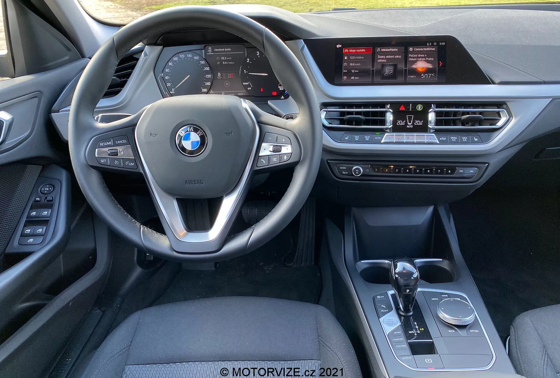 Vue intérieure de la BMW Série 1 (BMW 116i) 2019 depuis la perspective du siège passager avant, montrant le volant multifonction avec commandes intégrées, le tableau de bord numérique, le grand écran tactile d'info-divertissement, le système de contrôle de la climatisation, le levier de vitesses automatique, le sélecteur de mode de conduite et le bouton de contrôle iDrive.