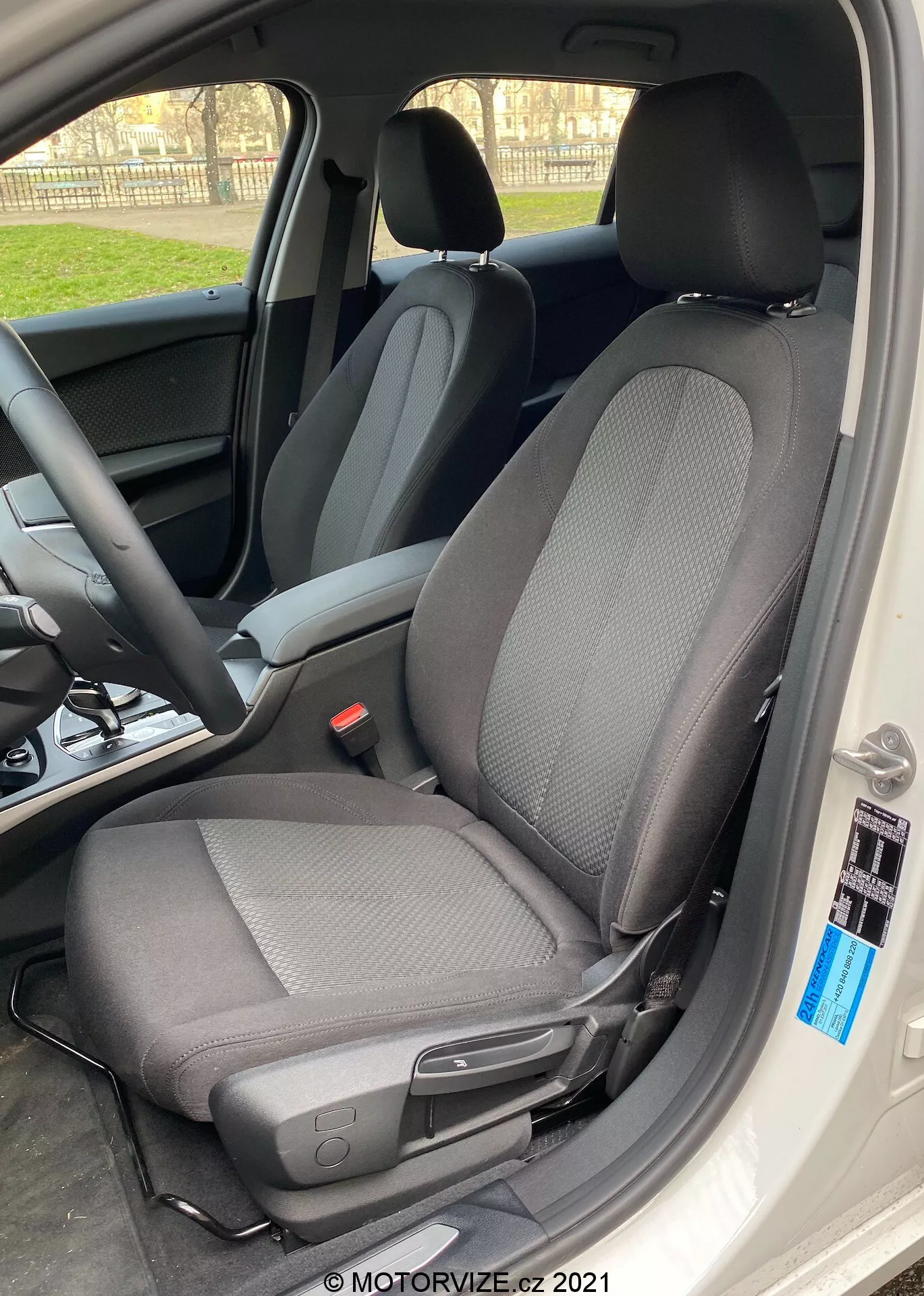 Gros plan de l'intérieur de la BMW 1er-Reihe 2019 (BMW-116i) avec un accent sur le siège du conducteur avec un design en tissu bicolore moderne, principalement noir avec des motifs gris clair, pris sous un angle latéral avec la porte de la voiture grande ouverte. Le siège est doté d'un appui-tête contre la lumière, créant un effet de silhouette autour des bords, et comprend un levier pour les réglages manuels vers l'avant ou l'arrière, ainsi qu'un petit bouton noir près du bord de l'intérieur de la voiture. La lumière naturelle du jour met en valeur la netteté du siège, avec un arrière-plan flou montrant le volant et le tableau de bord.