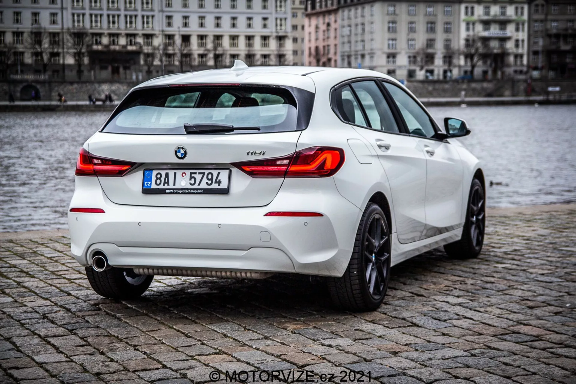 BMW Série 1 2019 (116i), modèle hatchback blanc, vue de trois quarts arrière avec des éléments détaillés tels que les feux arrière, le couvercle de coffre, le pare-chocs arrière, les pots d'échappement et une partie des roues arrière, stationné sur une surface pavée mouillée près d'un plan d'eau. L'image met en avant le spoiler arrière sportif, les clusters de feux arrière modernes et les badges visibles de BMW et de niveau de finition.
