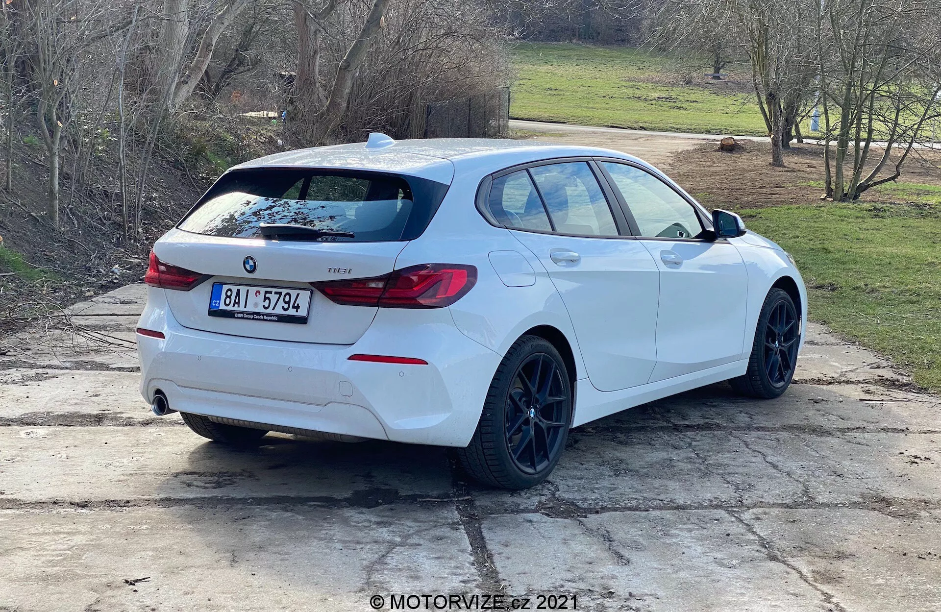 Vue arrière trois quarts d'une berline blanche BMW Série 1 (modèle BMW 116i 2019) garée en extérieur en plein jour. Le profil latéral et l'arrière de la voiture sont clairement visibles, avec l'emblème BMW, la désignation du modèle, les feux arrière distinctifs enveloppants, un aileron arrière, des jantes noires, un style de carrosserie type hatchback, des doubles tuyaux d'échappement, un essuie-glace arrière, des vitres légèrement teintées et des rétroviseurs avec clignotants intégrés. La voiture est positionnée sur une surface en béton avec des herbes et un arrière-plan rural ou suburbain.