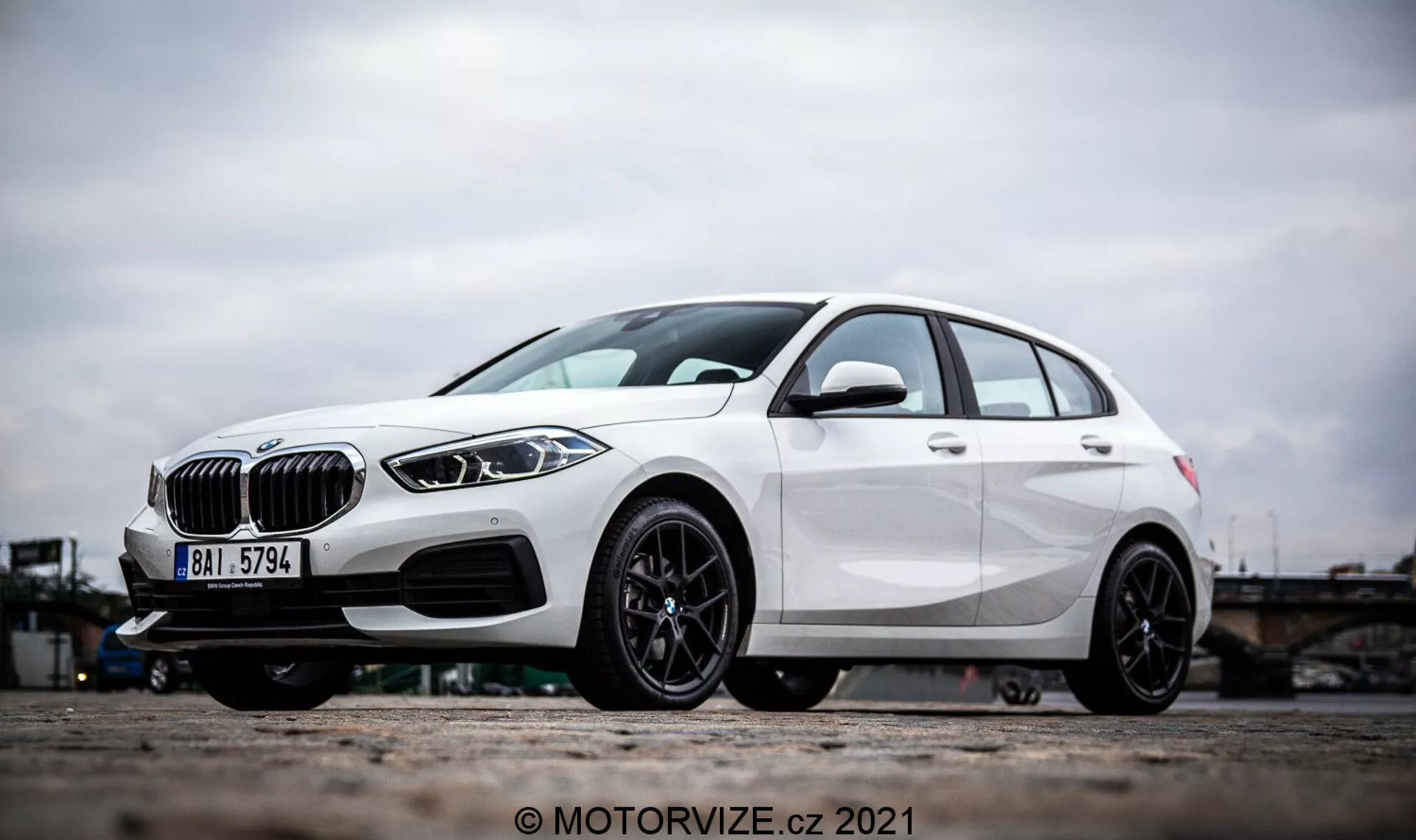 Vue latérale en angle d'une BMW Série 1 2019 (BMW 116i) blanche, présentant des éléments de design marquants comme une calandre en forme de rein, des phares LED élégants et un pare-chocs avant avec des prises d'air et des capteurs radar possibles pour l'assistance à la conduite. La photo se concentre sur les roues en alliage et la partie inférieure du véhicule, mettant en valeur le design sportif à plusieurs branches en alliage noir et le frein à disque performant. Les lignes de caractère et la sculpture aérodynamique de la berline soulignent davantage son extérieur moderne et dynamique.