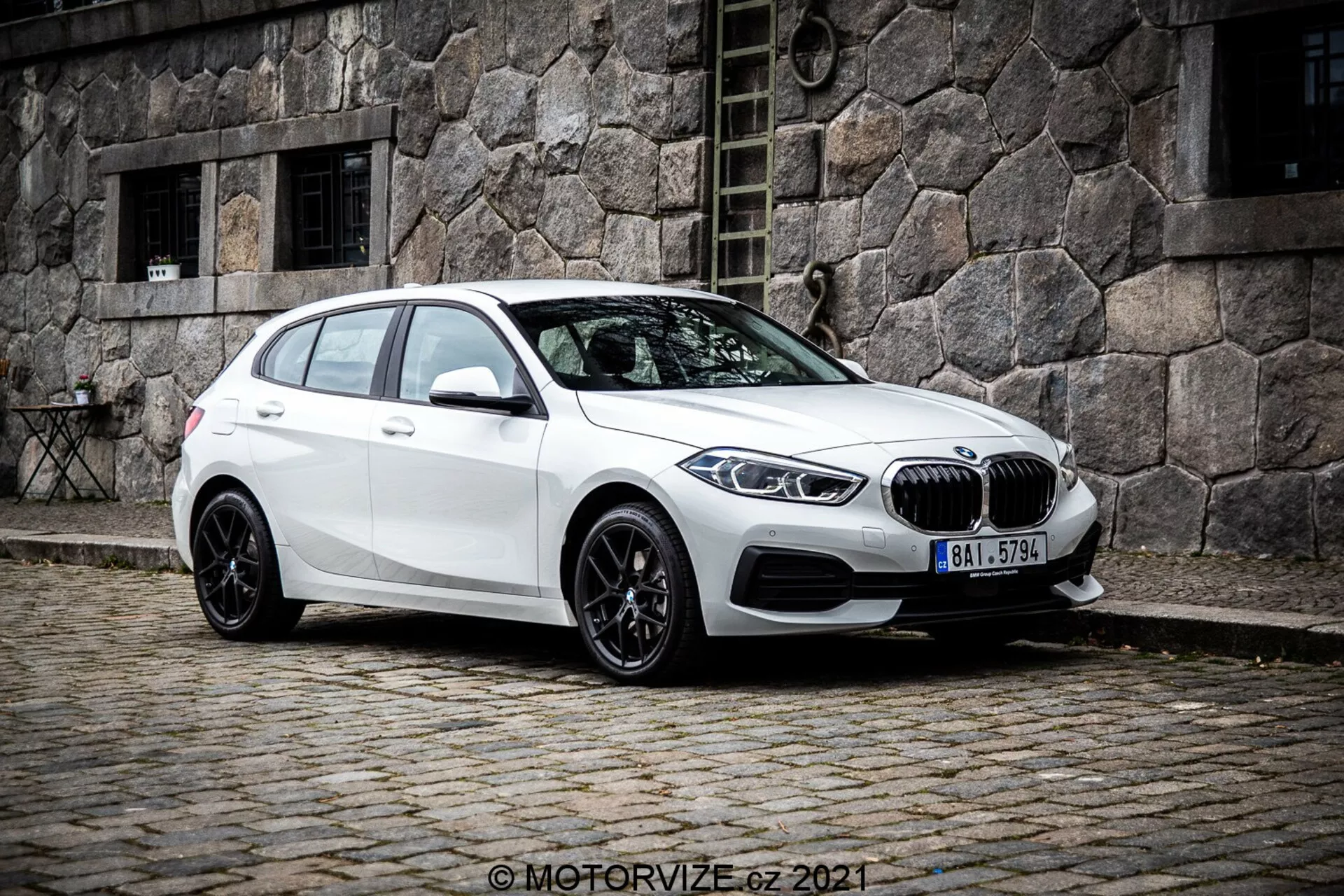 Une BMW 1er-Reihe 2019 (BMW 116i) blanche vue sous un angle frontal trois quarts, montrant le côté gauche du véhicule. La photo, prise d'un angle légèrement inférieur, accentue la posture et les lignes de design de la voiture. Elle est placée sur une rue pavée avec un vieux bâtiment en pierre en arrière-plan, créant une scène urbaine classique. La calandre en forme de rein et l'emblème rond de BMW sont bien visibles à l'avant, encadrés par des phares à LED angulaires élégants. La voiture est équipée de jantes en alliage noir à plusieurs rayons, d'une ligne de toit inclinée, de rétroviseurs latéraux couleur carrosserie avec clignotants intégrés et de fenêtres teintées pour les passagers arrière.