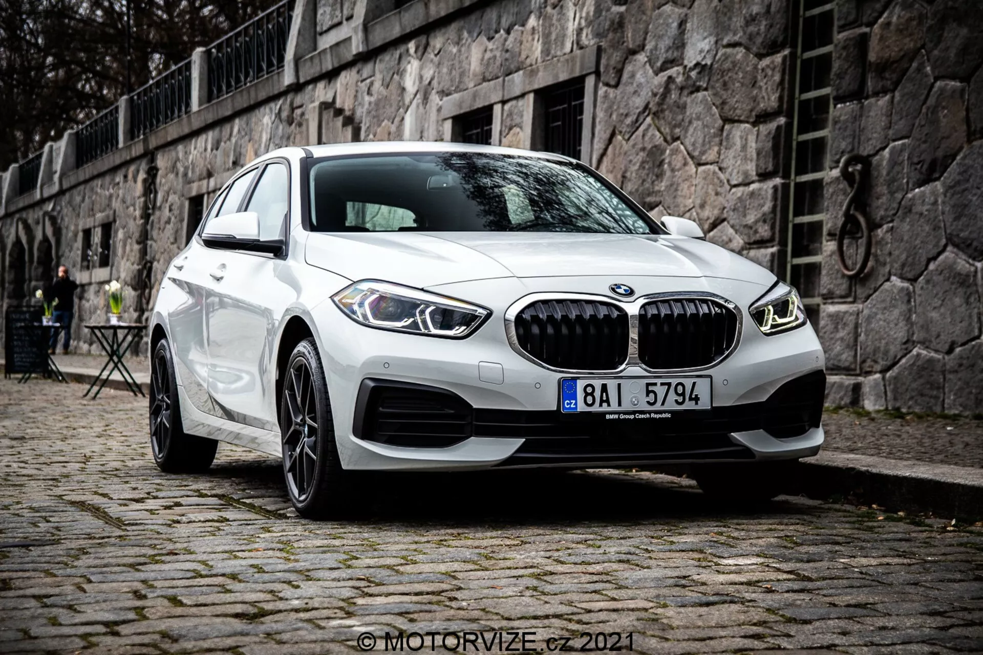 Modèle moderne de la BMW Série 1 blanche (BMW 116i) 2019 avec sa calandre caractéristique en forme de reins, ses feux de jour LED circulaires doubles et son design de pare-chocs avant sportif, stationné sur une surface en pavés dans un cadre européen historique avec un mur en pierre et des meubles de café en plein air en arrière-plan.