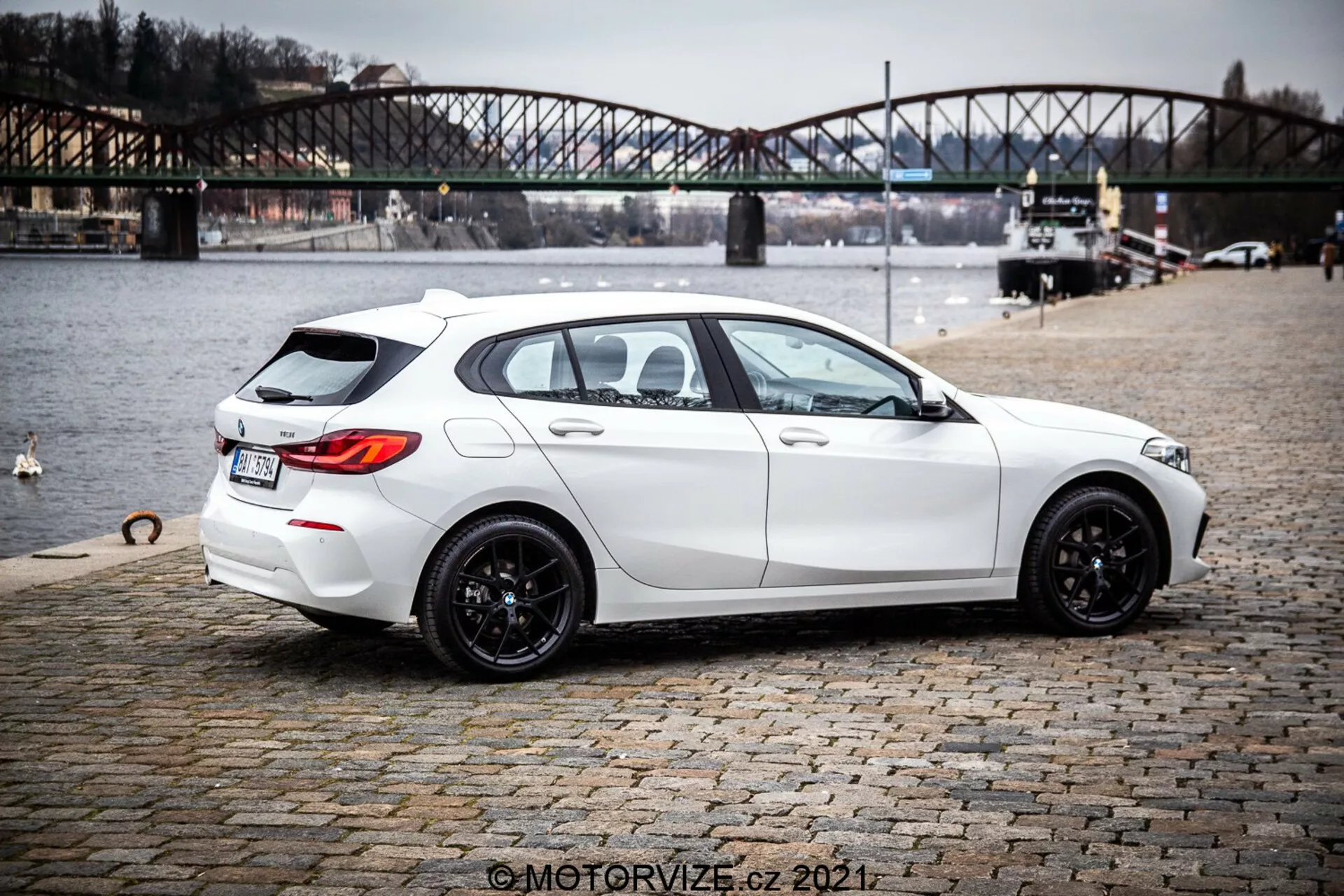 Image d'une berline blanche BMW 1er-Reihe 116i 2019 vue de trois quarts arrière, mettant en valeur son design moderne et élégant avec une allure sportive, des roues en alliage noir à plusieurs rayons, des fenêtres légèrement teintées et des fonctionnalités pratiques comme le dégivreur et l'essuie-glace de la lunette arrière. La voiture présente des lignes et des contours nets, un système d'échappement double, un subtil spoiler de toit arrière, une caméra de recul et des capteurs de stationnement. Elle est garée sur une surface pavée près d'un plan d'eau avec un pont urbain à l'arrière-plan, suggérant un contexte de style de vie en plein air.