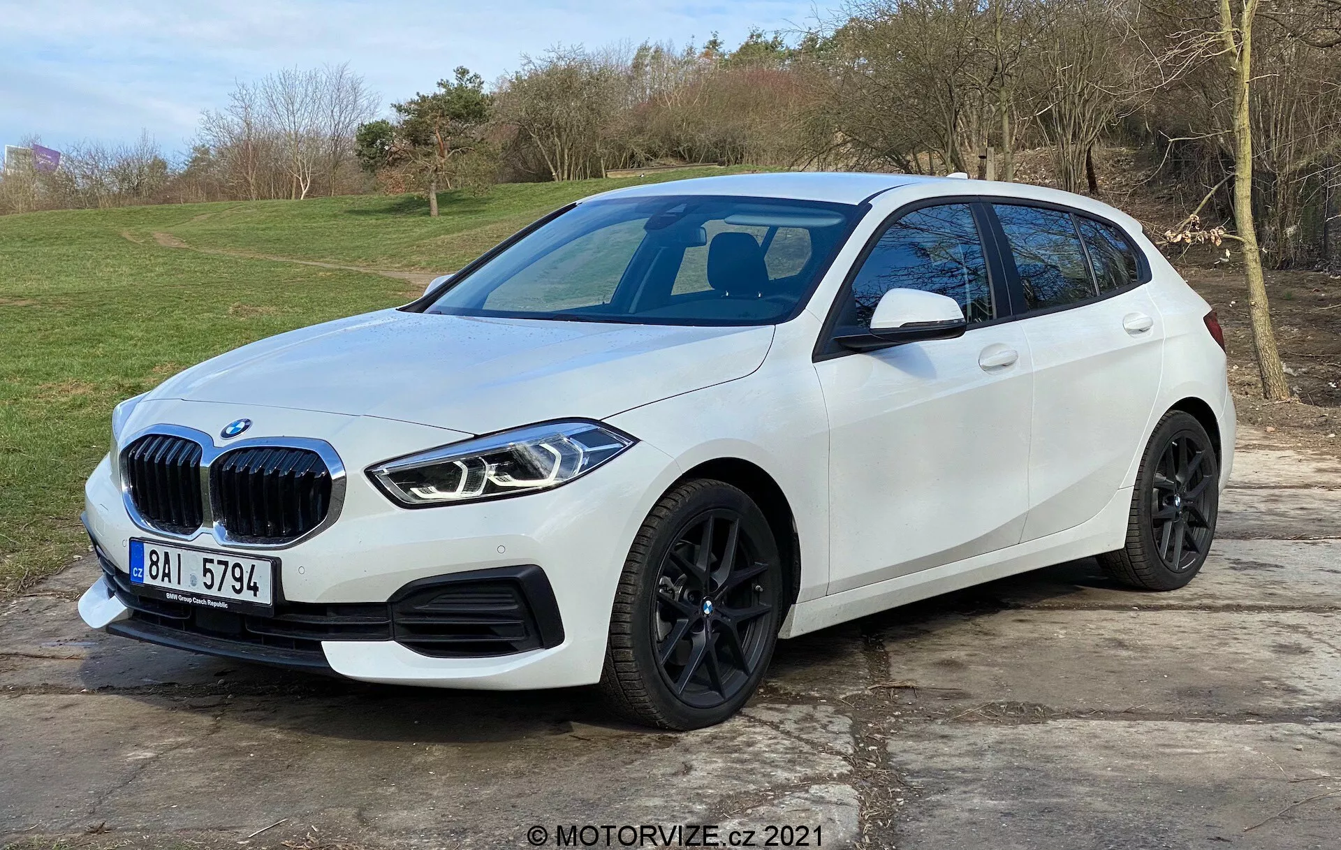 L'image montre une berline compacte BMW Série 1 blanche moderne (BMW 116i) positionnée en extérieur avec un arrière-plan naturel herbeux. Capturée d'une vue avant trois-quarts favorisant le côté conducteur, elle met en évidence la calandre distinctive du véhicule, les phares LED angulaires élégants, les jantes en alliage noires, le pli latéral bien défini et les indicateurs de clignotant intégrés sur les rétroviseurs. Le pare-chocs avant comporte des capteurs de stationnement, tandis qu'un toit panoramique en verre et une possible caméra avant peuvent être inférés. La voiture est stationnée sur une zone pavée et occupe la majeure partie du cadre, suggérant un design automobile moderne et des fonctionnalités technologiques avancées.