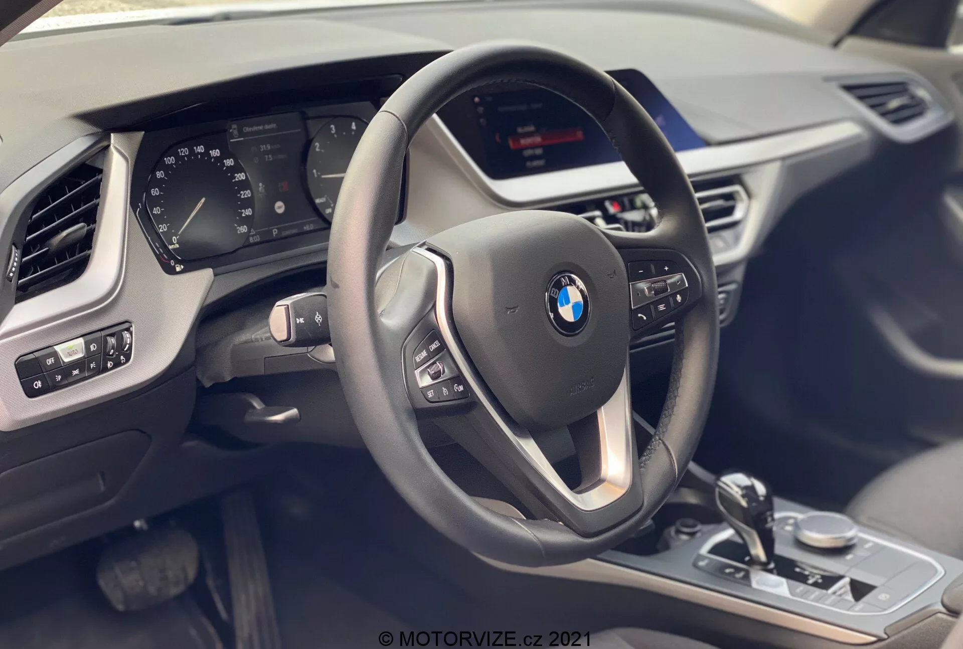 Vue intérieure de la BMW Série 1 2019 (BMW 116i) depuis le siège passager, mettant en avant le cockpit du conducteur avec un volant moderne à trois branches équipé de boutons multifonctions et recouvert de cuir avec des coutures contrastées. Derrière le volant, un tableau de bord numérique partiellement visible affiche diverses métriques du véhicule. La console centrale abrite un écran d'infodivertissement non visible et des commandes visibles telles que le sélecteur de vitesse, le bouton de commande iDrive, le sélecteur de mode de conduite et les commandes des capteurs de stationnement, avec des finitions métalliques et une garniture noire brillante. Le panneau de porte du conducteur montre les réglages des miroirs et les commandes des vitres électriques. Une partie de la grille d'aération latérale du passager et la texture du tableau de bord sont également visibles, soulignant le luxe, la technologie et le design moderne.