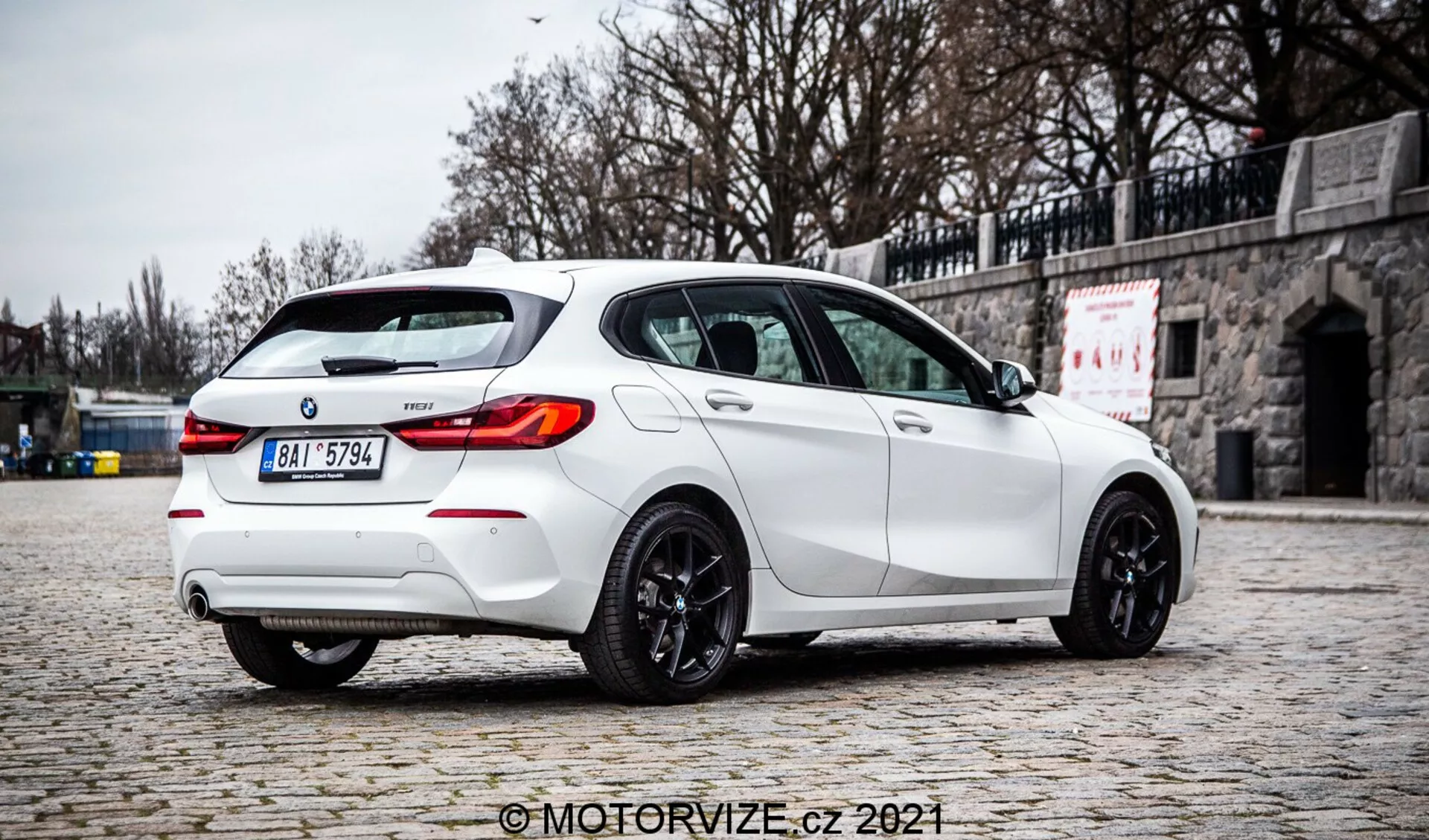 Photographie de haute qualité d'une BMW Série 1 2019 128ti hatchback blanche prise sous un angle trois-quarts arrière, mettant en évidence le côté droit. La voiture est garée sur un pavage en pierre avec un mur de pierre en arrière-plan, soulignant son design sportif avec un aileron de toit arrière, un système d'échappement double, un diffuseur arrière, des feux arrière à LED, des vitres teintées et des roues en alliage de couleur foncée. L'éclairage diffus, l'absence d'ombres dures et les détails clairs suggèrent un cadre nuageux, offrant une représentation authentique de la couleur et des caractéristiques du véhicule. L'image porte le filigrane 'MotorVize.cz 2021'.