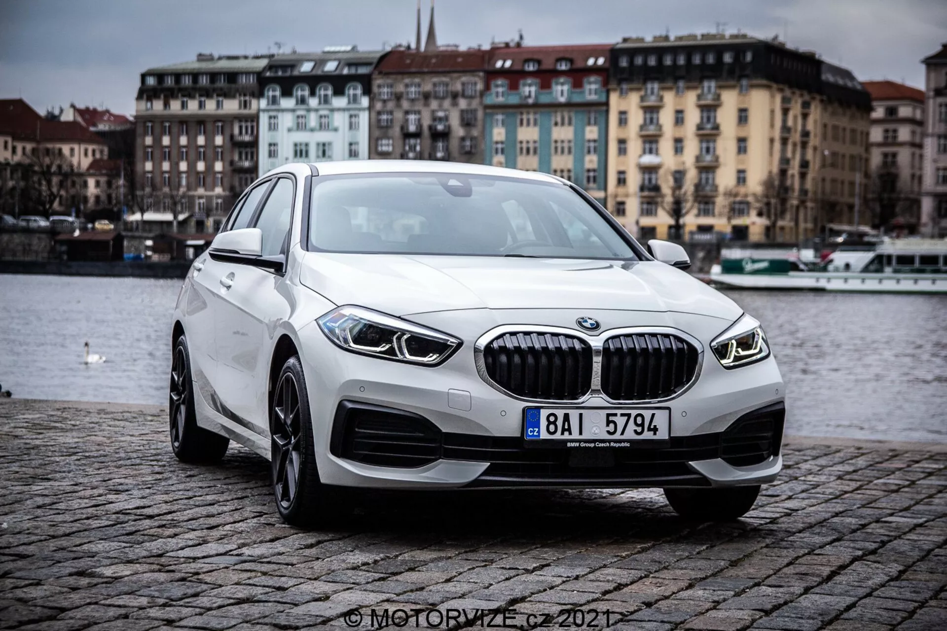 Image montrant une BMW 1er-Reihe 2019 (BMW 116i) vue de trois quarts avant, mettant en valeur les contours et les éléments de design de la berline de luxe moderne. La voiture est située à l'extérieur sur un remblai pavé près d'un plan d'eau, sous un ciel nuageux. Les caractéristiques marquantes incluent une grille avec des lamelles verticales, des phares LED élégants, des antibrouillards intégrés, des roues en alliage et un profil aérodynamique.