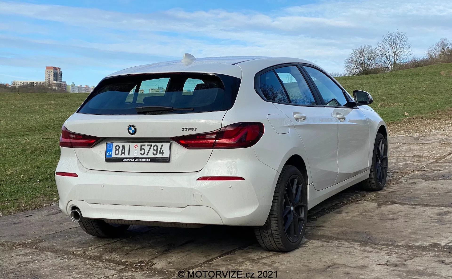 Vue arrière trois-quarts d'une BMW Série 1 118i blanche, garée sur un sol irrégulier. La voiture présente des feux arrière distinctifs en forme de 'L', un badge BMW et un badge modèle indiquant '118i'. Elle dispose d'un système d'échappement double avec un tuyau visible du côté droit du pare-chocs arrière. Le véhicule est équipé de roues en alliage noires à plusieurs branches et d'un essuie-glace arrière. Un spoiler de toit ajoute à l'aspect sportif et le profil latéral révèle des fenêtres légèrement teintées, des plis de carrosserie et des poignées de porte et rétroviseurs assortis. L'angle bas de la caméra met en évidence la posture de la voiture et le design des roues.