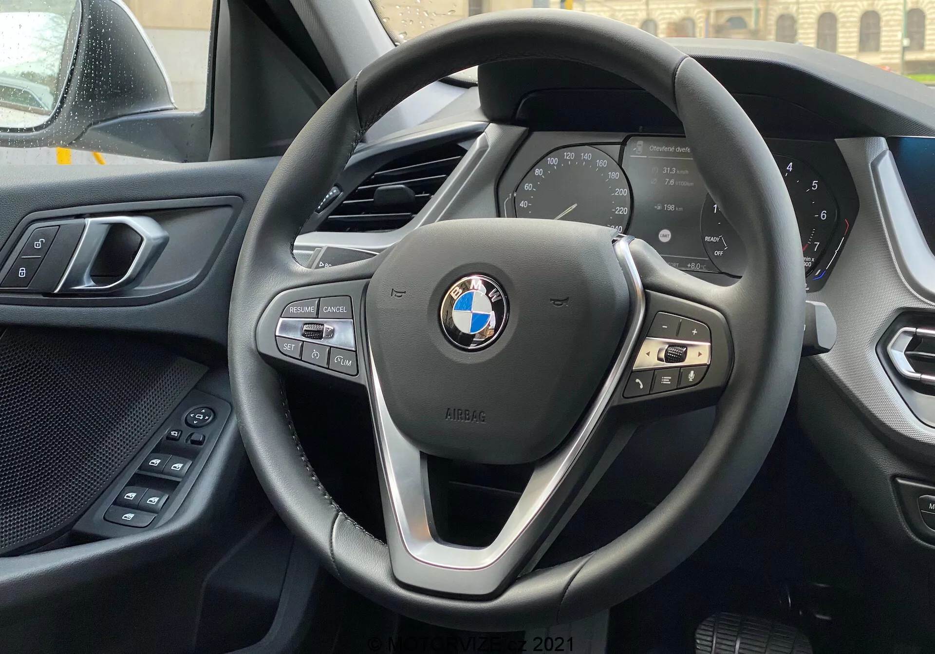 Vue intérieure d'une BMW Série 1 2019 (BMW 116i) depuis le siège du conducteur, axée sur le volant moderne à trois branches BMW avec des boutons de commande pour le multimédia, le téléphone et le régulateur de vitesse, et le tableau de bord numérique affichant les principales données du véhicule telles que la vitesse et les tr/min. La bordure supérieure de la console centrale est visible à l'arrière-plan, et une partie de la portière du conducteur avec des commandes électriques de confort est visible à gauche. Le tableau de bord et le panneau de porte présentent une combinaison de matériaux doux au toucher et de fabrication de qualité. L'emblème BMW et le badge iPERFORMANCE sont bien en évidence sur le volant, suggérant une technologie efficace ou hybride.