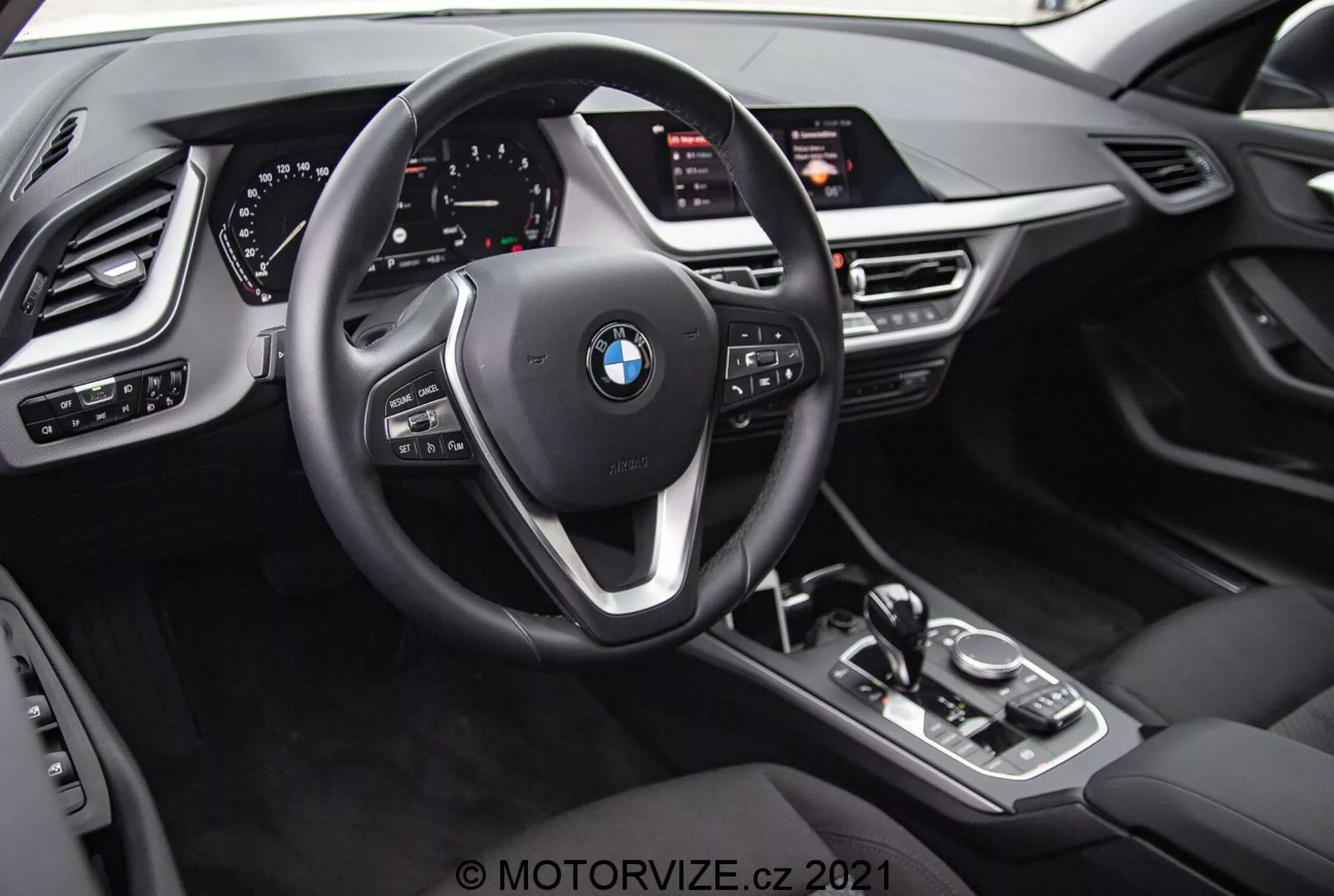 Vue intérieure de la BMW 1er-reihe 2019 (BMW 116i) du point de vue du conducteur. L'image met en avant l'emblème BMW sur le volant à trois branches avec des boutons multifonctions et des palettes de changement derrière le volant. Derrière le volant se trouve un tableau de bord entièrement numérique avec des cadrans illuminés et divers affichages informatifs. La console centrale comporte un levier de sélection de boîte de vitesses automatique, un bouton de contrôle iDrive, des boutons pour les modes de conduite et un grand écran d'infodivertissement orienté vers le conducteur. Le design intérieur comprend des textures similaires au cuir, des accents métalliques et des commandes supplémentaires pour les phares, les fenêtres et les réglages des rétroviseurs avec des fonctions de mémoire pour le siège du conducteur.