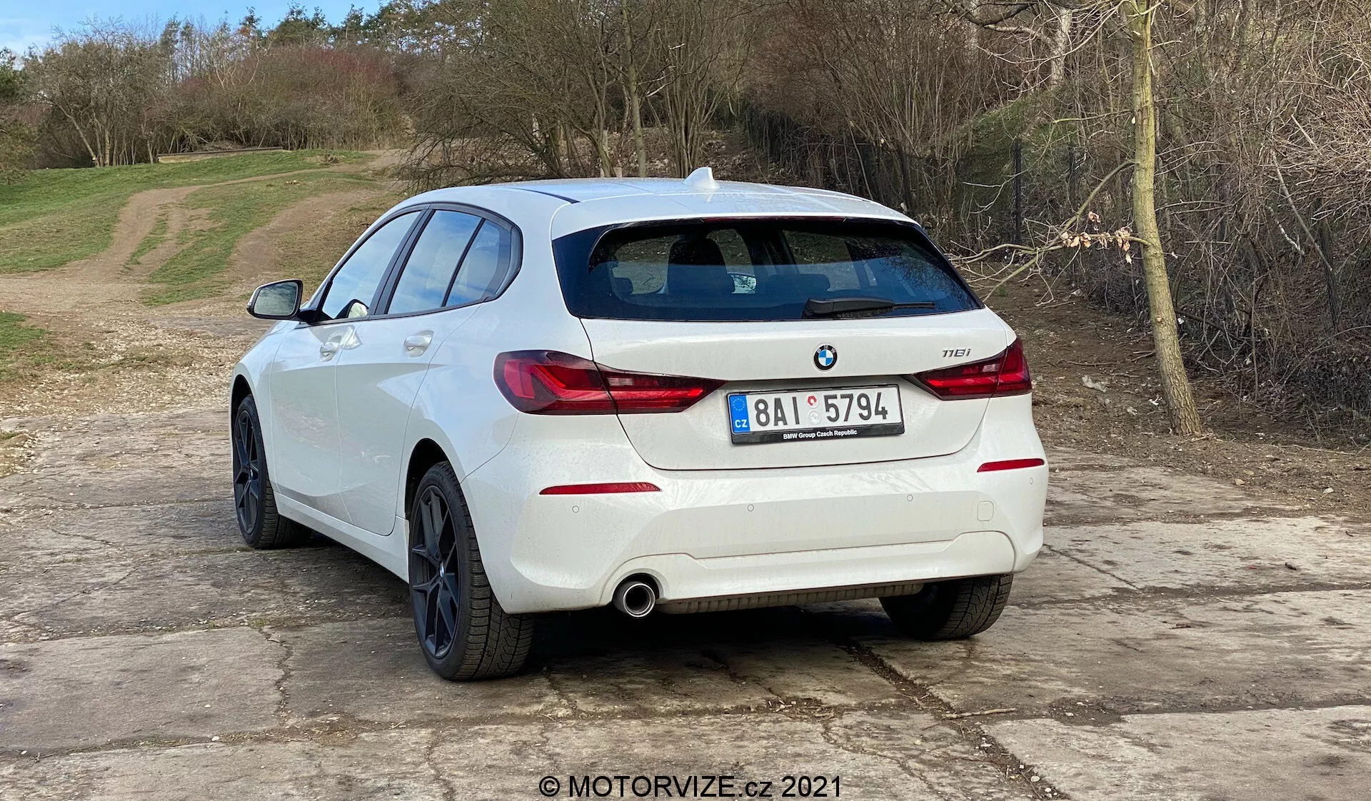 L'image montre une BMW 1er-reihe 2019 (BMW 116i) blanche, vue sous un angle arrière trois quarts. L'arrière du véhicule, y compris les feux arrière horizontaux, le système d'échappement double et les capteurs de stationnement intégrés dans le pare-chocs arrière, est clairement visible. L'emblème BMW et le badge du modèle sont sur le hayon. La voiture est située sur une surface de gravier avec un cadre naturel et rural. L'image suggère des thèmes automobiles tels que le design des véhicules, les modèles contemporains, le contenu spécifique à BMW, les systèmes d'échappement et la technologie d'assistance au stationnement. La marque d'eau MOTORVIZE.cz 2021 indique une origine dans une revue ou publication automobile.