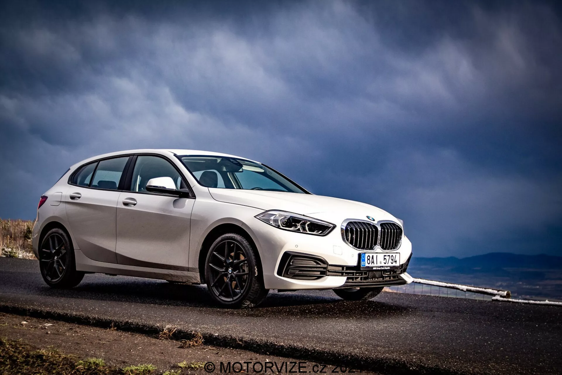 BMW Série 1 2019 (BMW-116i) capturée en vue avant trois quarts, présentant son profil avant et latéral. La voiture, finie en blanc brillant avec des accents noirs contrastants comme les coques de rétroviseurs, les garnitures de fenêtres et les roues à plusieurs branches, est positionnée sur le côté gauche du cadre contre un fond de route asphaltée s'étendant au loin et de ciel orageux. Les caractéristiques visibles comprennent des phares détaillés avec des éléments circulaires, une calandre en forme de rein proéminente, un pare-chocs avant avec des évents d'admission d'air et des capteurs de stationnement potentiels.