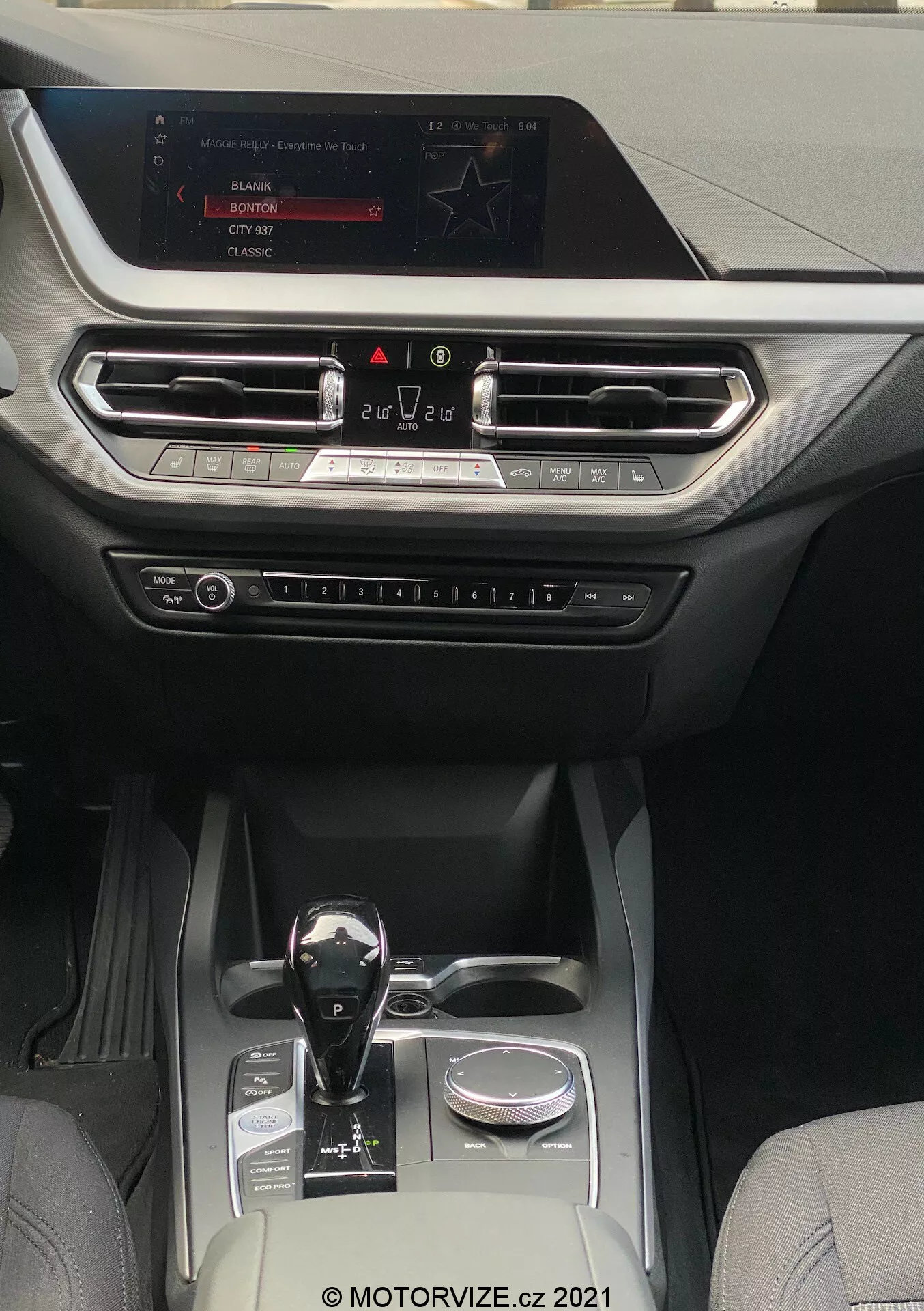 Vue intérieure d'une BMW 1er-reihe 2019 (BMW-116i) mettant en valeur un habitacle moderne et luxueux axé sur la console centrale et la zone du tableau de bord. La perspective est depuis la zone des sièges arrière, regardant vers l'avant vers l'écran d'infodivertissement monté en haut du tableau de bord. L'écran affiche des options pour la radio, les médias et les réglages de la voiture. Sous l'écran, des bouches d'aération alignées horizontalement avec des accents métalliques et une garniture décorative s'étendent sur toute la largeur du tableau de bord. Sous les bouches d'aération, des boutons et des commandes physiques pour le contrôle du climat et d'autres réglages sont visibles. Le levier de vitesses automatique présente une finition noire brillante avec des accents métalliques, positionné à côté d'une molette rotative. La console centrale comprend deux porte-gobelets, un petit compartiment de rangement et un élément décoratif argenté, indiquant un habitacle bien aménagé combinant du cuir et des tissus synthétiques de haute qualité, adapté aux catégories de véhicules de milieu à haut de gamme axées sur la technologie et le confort.