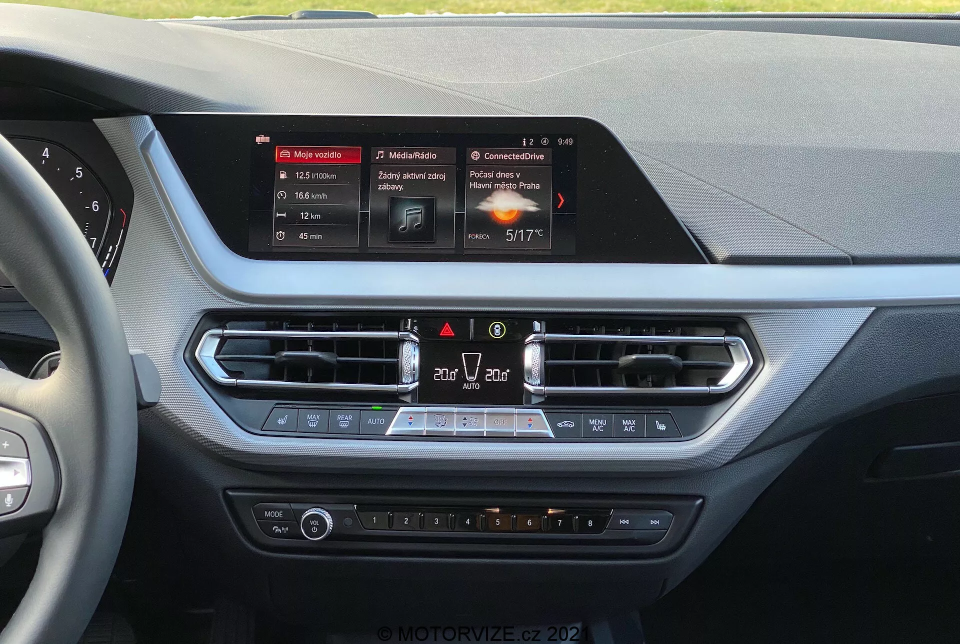 Vue intérieure d'une BMW série 1 de 2019 (BMW-116i) depuis la perspective du conducteur, mettant en avant la console centrale du tableau de bord. Au centre se trouve l'écran tactile rectangulaire et illuminé du système d'infodivertissement, affichant la navigation, les médias et les détails climatiques. Sous l'écran se trouvent des bouches d'aération avec garniture métallique, des commandes d'urgence et de recirculation d'air, suivies d'un panneau de contrôle climatique avec boutons de température, vitesse du ventilateur et sièges chauffants. Les matériaux présentent des surfaces douces au toucher et des accents chromés, suggérant une focalisation sur le confort et la commodité du conducteur et du passager avant.
