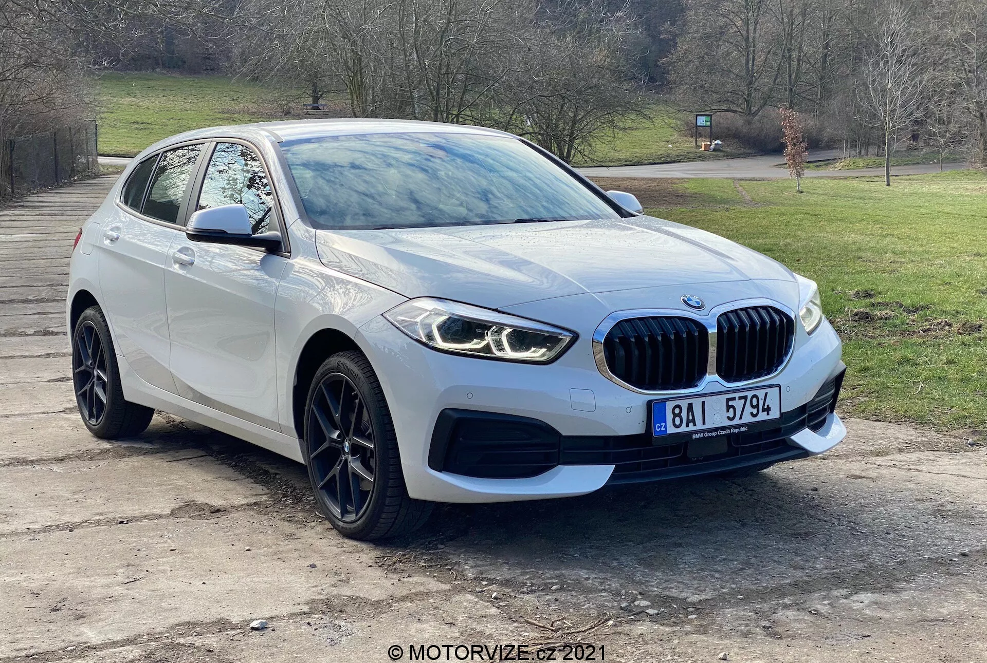 Véhicule moderne BMW 116i de la Série 1 de 2019 photographié sous un angle de trois quarts avant, montrant la calandre avant avec emblème, des phares LED élégants, feux de brouillard, pare-chocs avant et les lignes aérodynamiques du capot menant au pare-brise. La roue et l'aile côté conducteur, avec le rétroviseur latéral équipé de clignotants intégrés, mettent en avant le design avancé et la technologie des capteurs de stationnement du véhicule. Photographié en extérieur avec un arrière-plan d'arbres et d'herbes, la voiture dispose de jantes en alliage et d'une calandre caractéristique en forme de rein, indiquant le luxe et la performance.