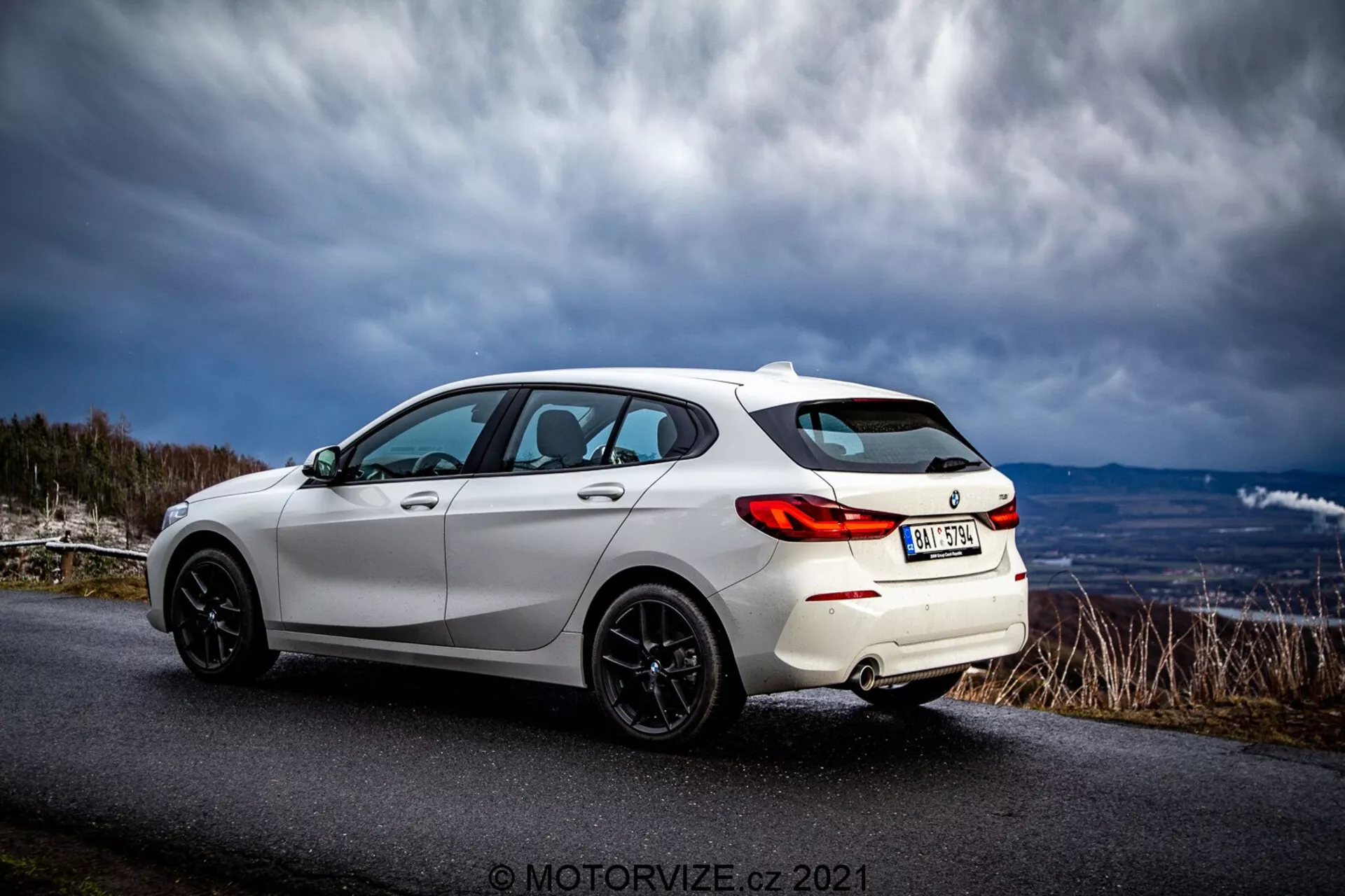 Un hatchback blanc moderne BMW 116i 2019 capturé en vue arrière trois quarts. Les caractéristiques sportives de l'extérieur comprennent des feux arrière à LED, des jantes en alliage noir à rayons multiples, un spoiler arrière, des vitres teintées et des lignes de carrosserie élégantes qui soulignent son design aérodynamique. Le véhicule est garé sur une route asphaltée dans une zone rurale ou montagneuse sous un ciel dramatique.