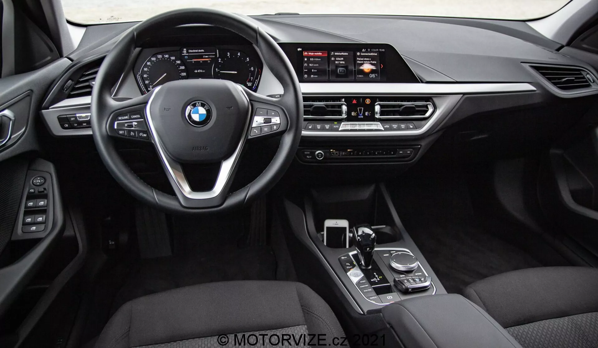 Vue intérieure d'une BMW Série 1 2019 (BMW 116i) prise depuis les sièges arrière, montrant le tableau de bord complet, le volant avec le logo BMW et les boutons de commande, et la console centrale. Les éléments visibles incluent l'écran large d'infodivertissement, les contrôles manuels du climat, les bouches d'aération, le levier de vitesses, le cadran rotatif et le pavé tactile. L'intérieur est fini en cuir noir ou simili-cuir avec des éléments de finition métalliques et brillants, mettant en valeur le luxe et la technologie d'interface avancée du conducteur.
