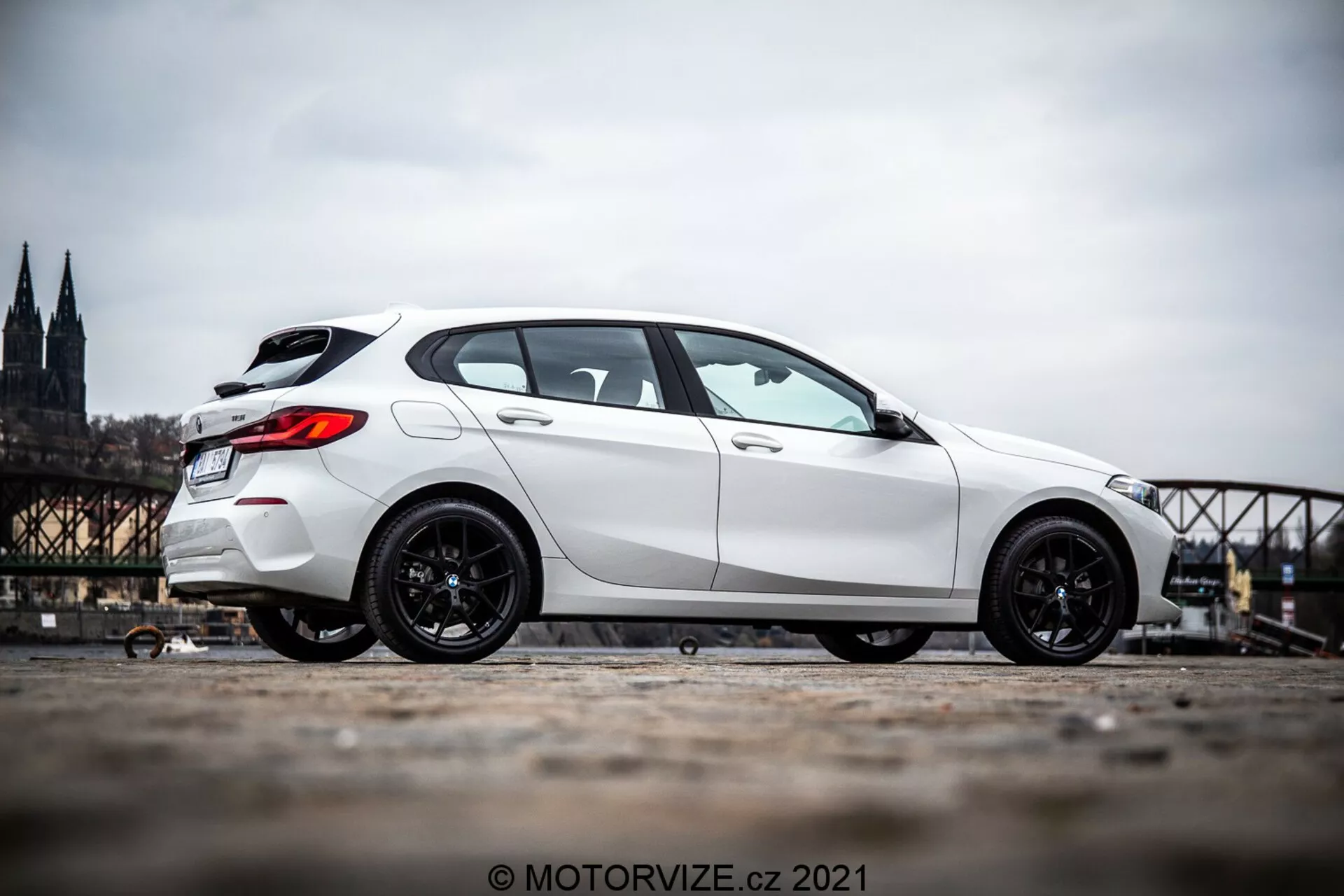 Une BMW Série 1 (BMW 116i) blanche de 2019 photographiée sous un angle latéral de profil, montrant le côté du véhicule depuis juste avant la roue avant jusqu'à l'arrière. La voiture est positionnée sur le côté gauche de l'image avec un arrière-plan urbain historique flou qui inclut une cathédrale et un pont. L'image met en évidence les jantes sportives sombres de la voiture et sa posture dominante grâce à la perspective en contrebas et à l'inclinaison vers le haut de la caméra. La photo est prise en extérieur pendant la journée, révélant les détails esthétiques de la voiture à la lumière naturelle du jour.