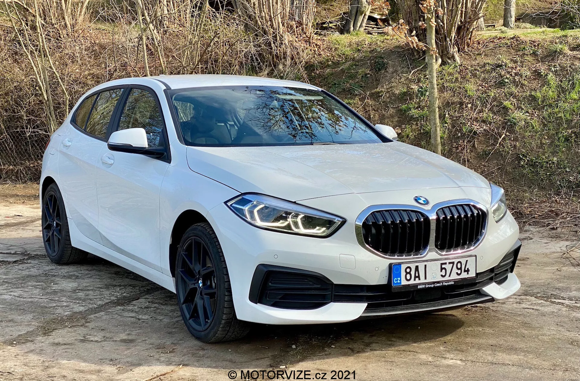 BMW 116i Série 1 de 2019 en blanc, capturé sous un angle de trois quarts avant, mettant en valeur sa grille avant en forme de rein proéminente, ses phares LED angulaires, ses élégantes roues noires avec des pneus à profil bas et ses lignes aérodynamiques. Positionné en extérieur sur une surface plane avec de la végétation naturelle en arrière-plan, la prise de vue met en avant le design moderne et les caractéristiques sportives de la voiture, y compris les capteurs de stationnement et les rétroviseurs latéraux avec clignotants intégrés.