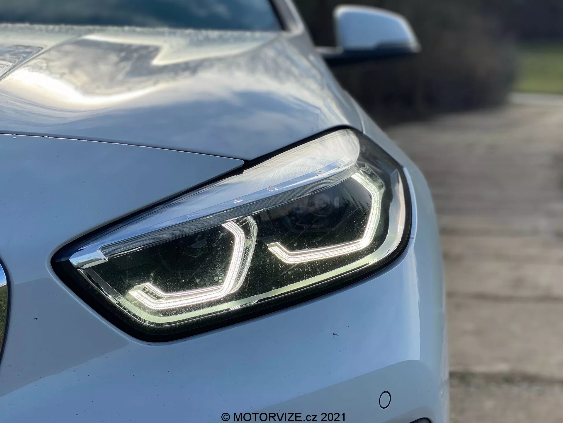 Gros plan sur la BMW Série 1 2019 (BMW 116i) sous un angle de quart avant, mettant en évidence le côté gauche et se concentrant sur le design moderne et élégant du phare avant gauche avec des feux de jour à LED et un couvercle de lentille transparent. Les lignes aérodynamiques du capot et de l'aile sont partiellement visibles, montrant la couleur métallique claire et la finition brillante de la voiture. Un arrière-plan flou met en avant les caractéristiques technologiques avancées du phare, suggérant des lampes LED ou HID, éventuellement équipées de la technologie d'éclairage adaptatif.