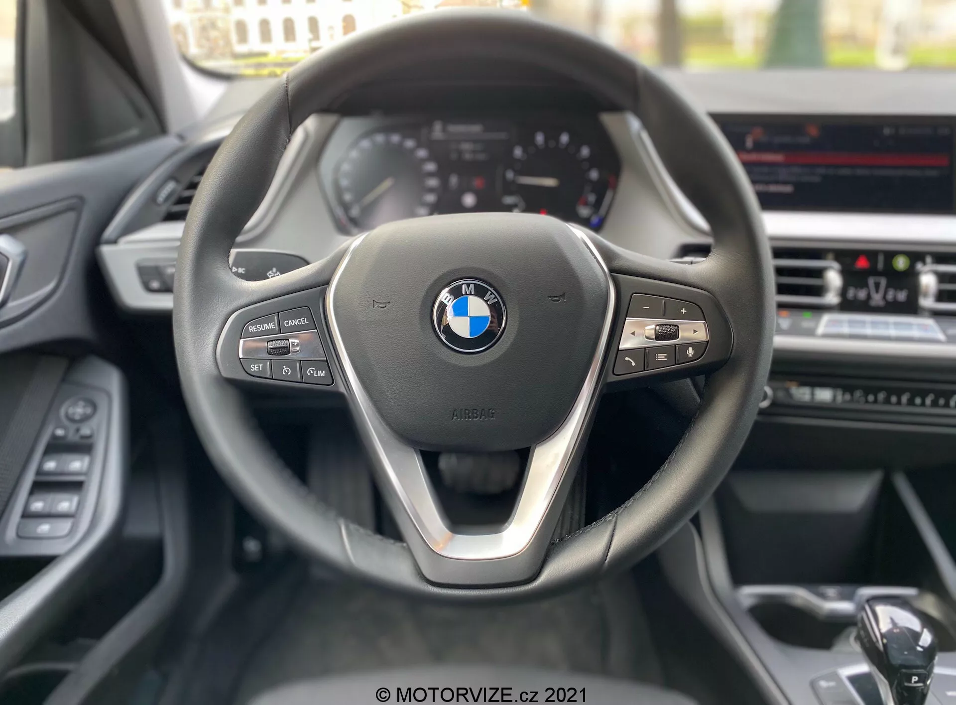 Perspective du siège conducteur d'une BMW Série 1 de 2019 (BMW-116i), centrée sur le volant contemporain à trois branches avec un accent métallique sur la branche inférieure et le logo BMW bien visible au centre. Le côté gauche du volant comporte des commandes de médias et de communication, tandis que le côté droit comprend des boutons de régulateur de vitesse et une palette de changement de vitesse visible. En arrière-plan, un tableau de bord partiellement visible avec des jauges de style analogique et un écran d'infodivertissement sur la console centrale suggèrent des fonctionnalités avancées telles que la navigation GPS et le contrôle de la climatisation.