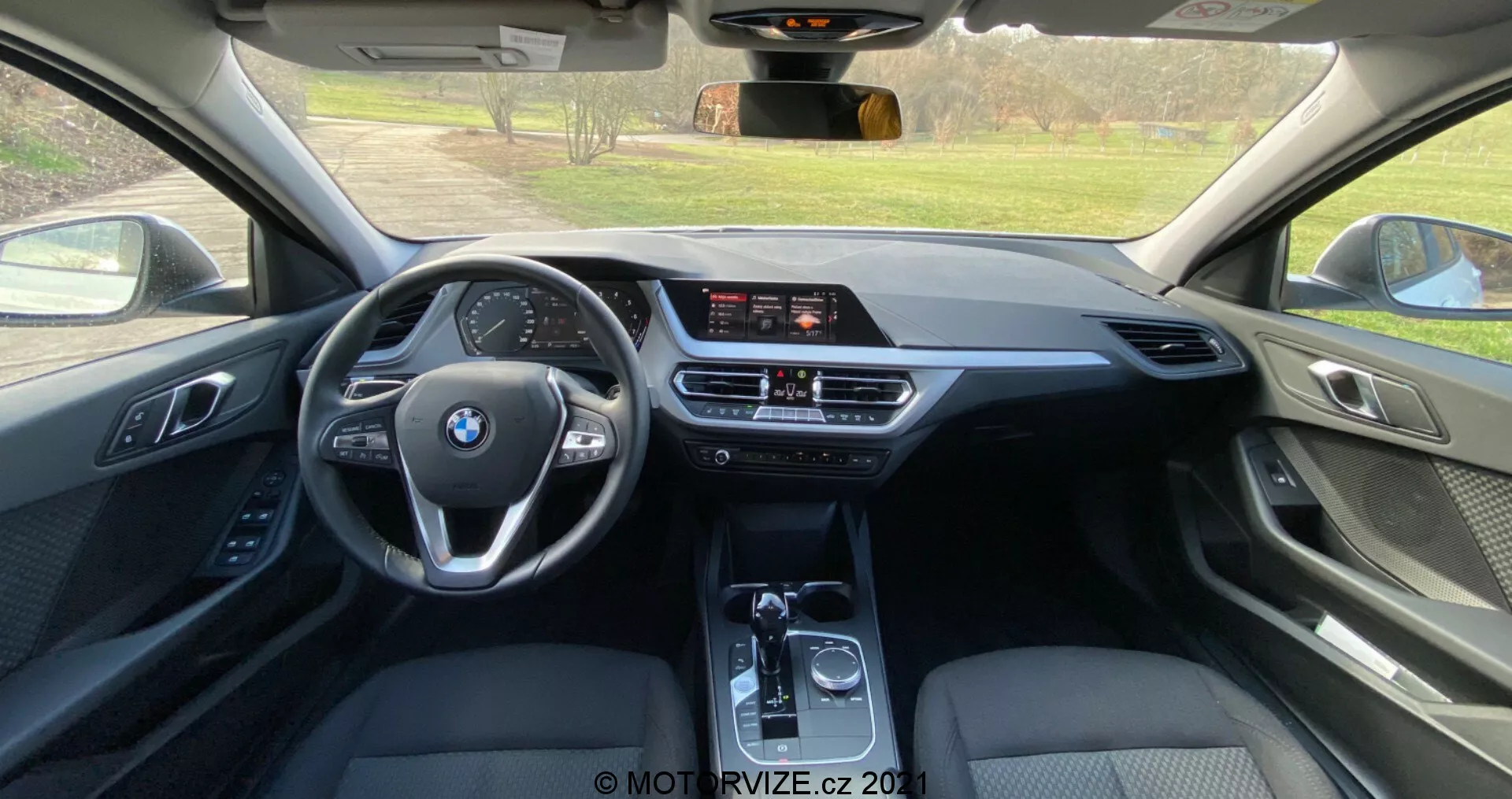 Vue intérieure depuis le siège arrière d'une BMW Série 1 2019 (BMW-116i), montrant un habitacle moderne avec un volant multifonction à trois branches portant le logo BMW, un écran d'info-divertissement autonome, une climatisation bi-zone et un levier de vitesse automatique. L'utilisation de cuir, de plastiques souples et d'accents métalliques met en évidence un niveau de finition haut de gamme.