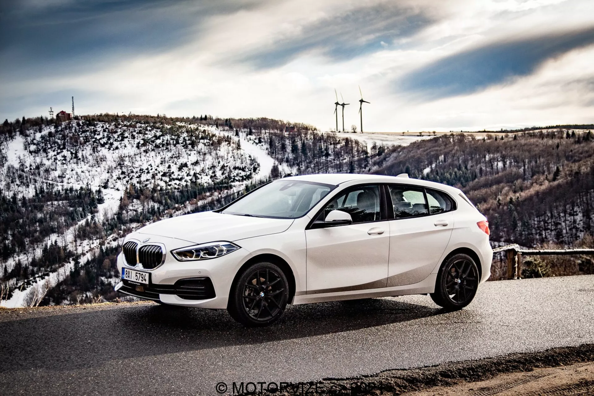 L'image montre une BMW 116i hatchback de 2019 blanche garée sur un point de vue pittoresque ou une route de montagne, vue sous un angle latéral avant mettant en évidence la calandre, les phares à LED, la roue avant, le profil latéral et un aperçu de l'arrière. La perspective est légèrement surélevée, mettant en valeur le toit de la voiture décalé sur la gauche, avec en arrière-plan des collines enneigées et des éoliennes lointaines. Le véhicule présente une calandre en forme de rognon, des pneus à profil bas sur des jantes en alliage à plusieurs branches, et des jantes de couleur foncée, des rétroviseurs latéraux avec clignotants intégrés et des vitres teintées. L'environnement suggère un temps clair, améliorant le caractère promotionnel et le côté visuellement attrayant de la photographie, potentiellement prise lors d'un road trip ou d'une revue automobile.