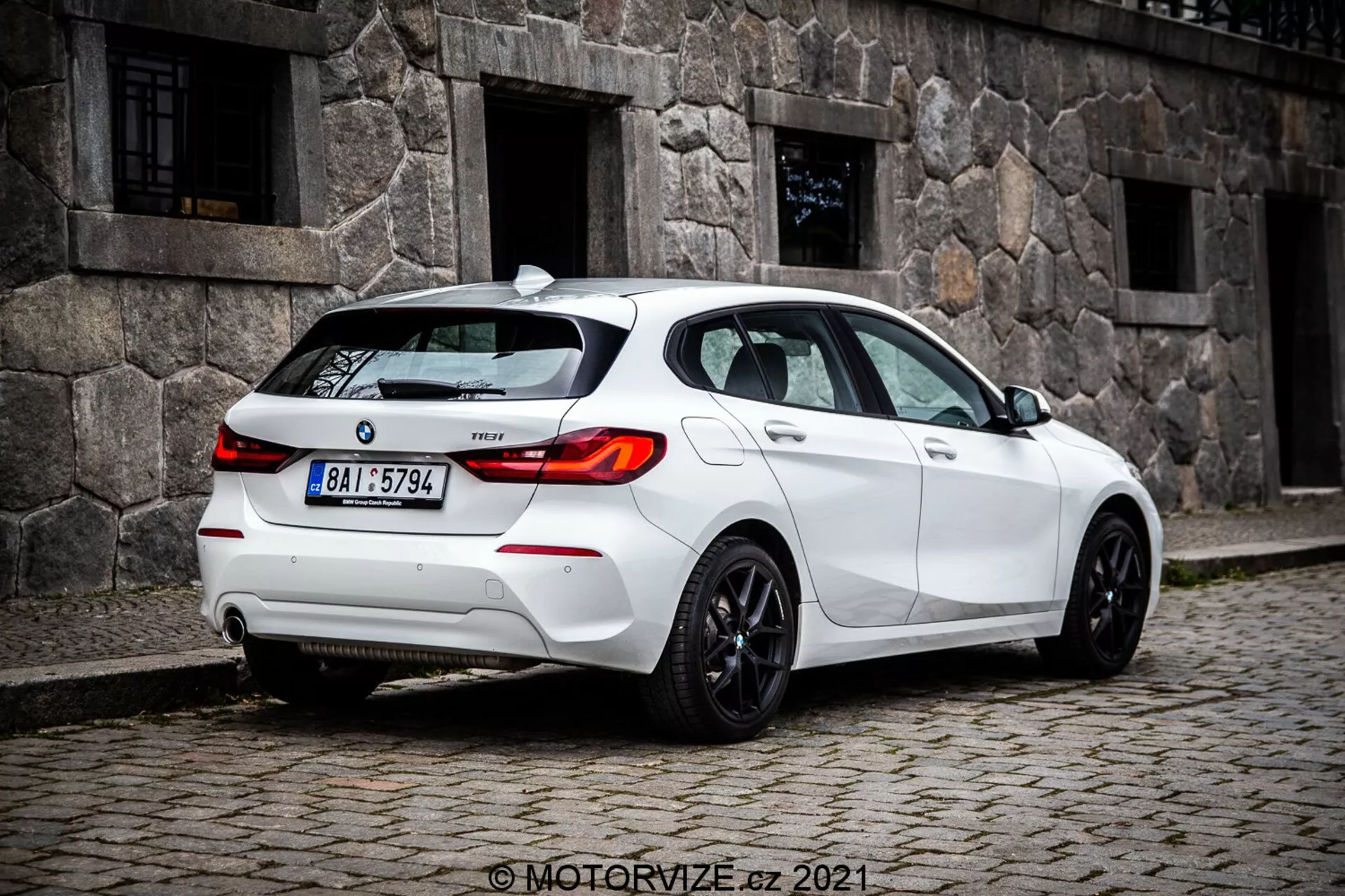 Image à mi-distance et au niveau du sol d'une berline compacte blanche BMW 1er-série 116i 2019 capturée sous un angle arrière de trois quarts, montrant en évidence les feux arrière au design LED, la lunette arrière et les jantes sportives. Les caractéristiques visibles incluent des capteurs sur le pare-chocs arrière probablement pour l'assistance au stationnement, une petite antenne sur le toit probablement pour GPS ou radio satellite, l'insigne du modèle sur le coffre, un système d'échappement double suggérant la performance, et les fenêtres teintées pour la confidentialité ou le confort.