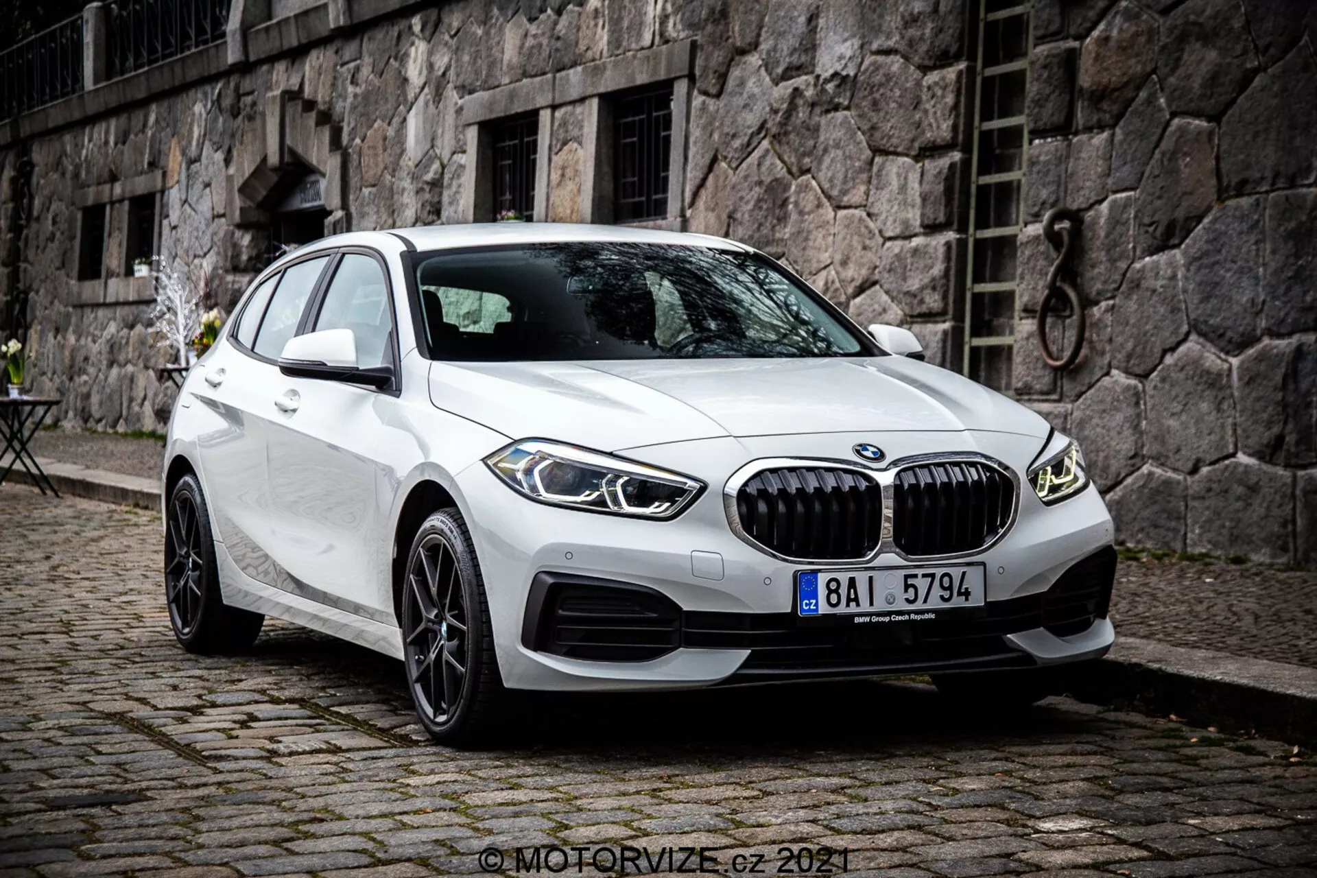 L'image montre une BMW Série 1 de 2019, spécifiquement la BMW 116i, de couleur blanche. Photographiée sous un angle de trois quarts avant, elle montre la partie avant de la voiture, le côté passager et le design détaillé de ses roues. La photo est prise d'un point de vue légèrement élevé, suggérant que le photographe était debout ou utilisait un trépied. La voiture est stationnée avec les roues avant tournées à droite, révélant clairement le design des jantes et le profil latéral du pneu. Les caractéristiques incluent une calandre avant proéminente avec des lamelles verticales, des lignes sculptées sur le capot et les panneaux latéraux, des phares LED fins et des prises d'air sur le pare-chocs pouvant contenir des capteurs de stationnement. Les rétroviseurs latéraux avec feux indicateurs intégrés et les fenêtres sans cadre soulignent son esthétique premium. Les jantes en alliage multi-branches suggèrent un niveau de finition supérieur ou une option améliorée. La lunette arrière fortement inclinée accentue le profil de la berline. Le véhicule est stationné sur un pavé de pierre dans un environnement urbain ou historique avec un bâtiment en pierre et une petite table de style bistrot en arrière-plan. L'image est nette et détaillée, capturée sous une lumière naturelle et nuageuse.