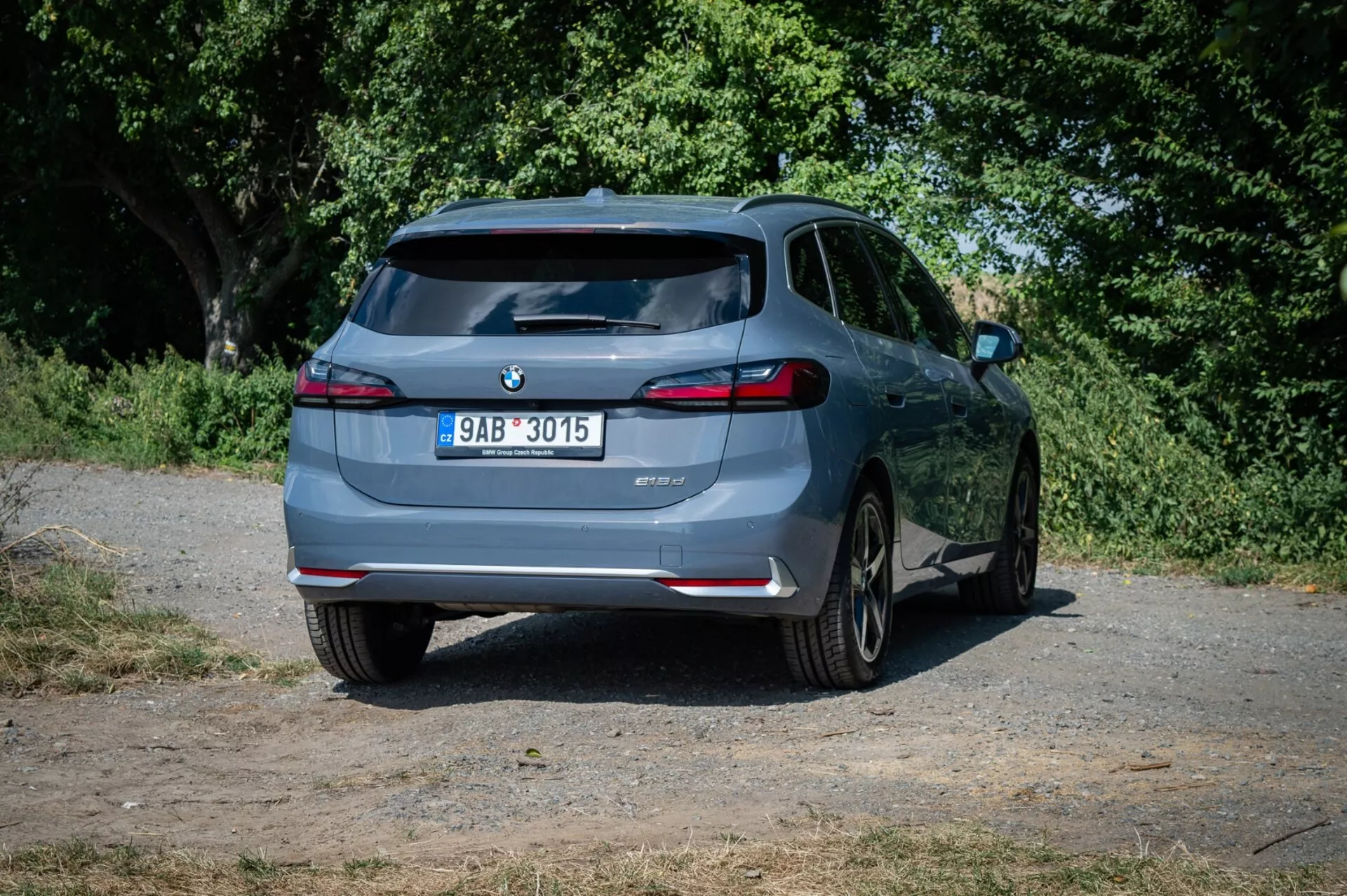 Hatchback BMW Série 2 Active Tourer 2022, vu d’un angle arrière légèrement décentré sur une route de campagne, mettant en avant son design arrière, feux arrière LED, double échappement et technologie d’assistance au stationnement dans un cadre naturel