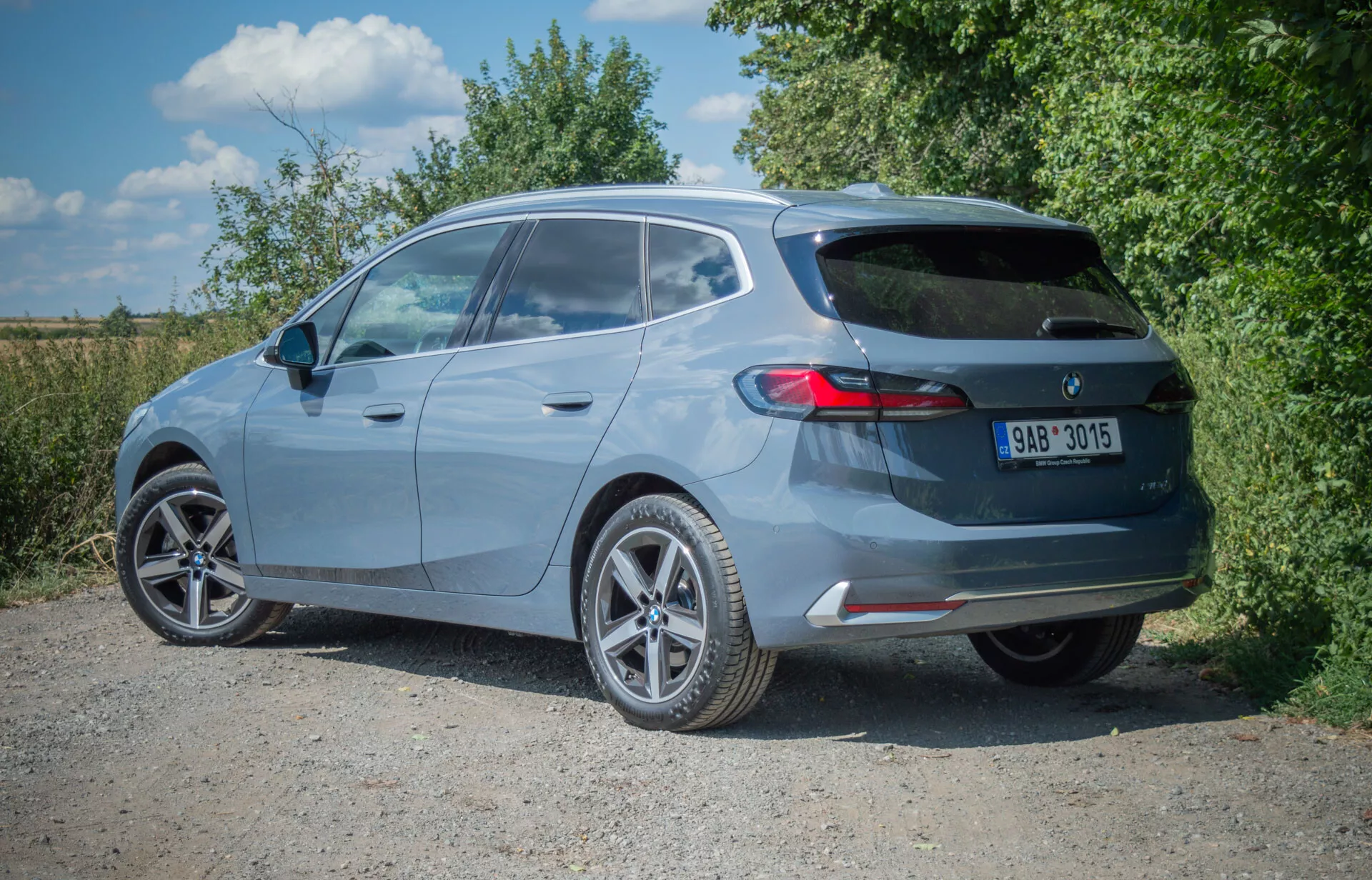 BMW Série 2 Active Tourer hatchback 2022 en peinture métallisée capturée depuis une vue arrière trois quarts, montrant des roues en alliage, aileron arrière, essuie-glace arrière, troisième feu stop, feux arrière horizontaux, échappement unique et pare-chocs arrière avec réflecteurs intégrés, garée sur une surface non pavée.
