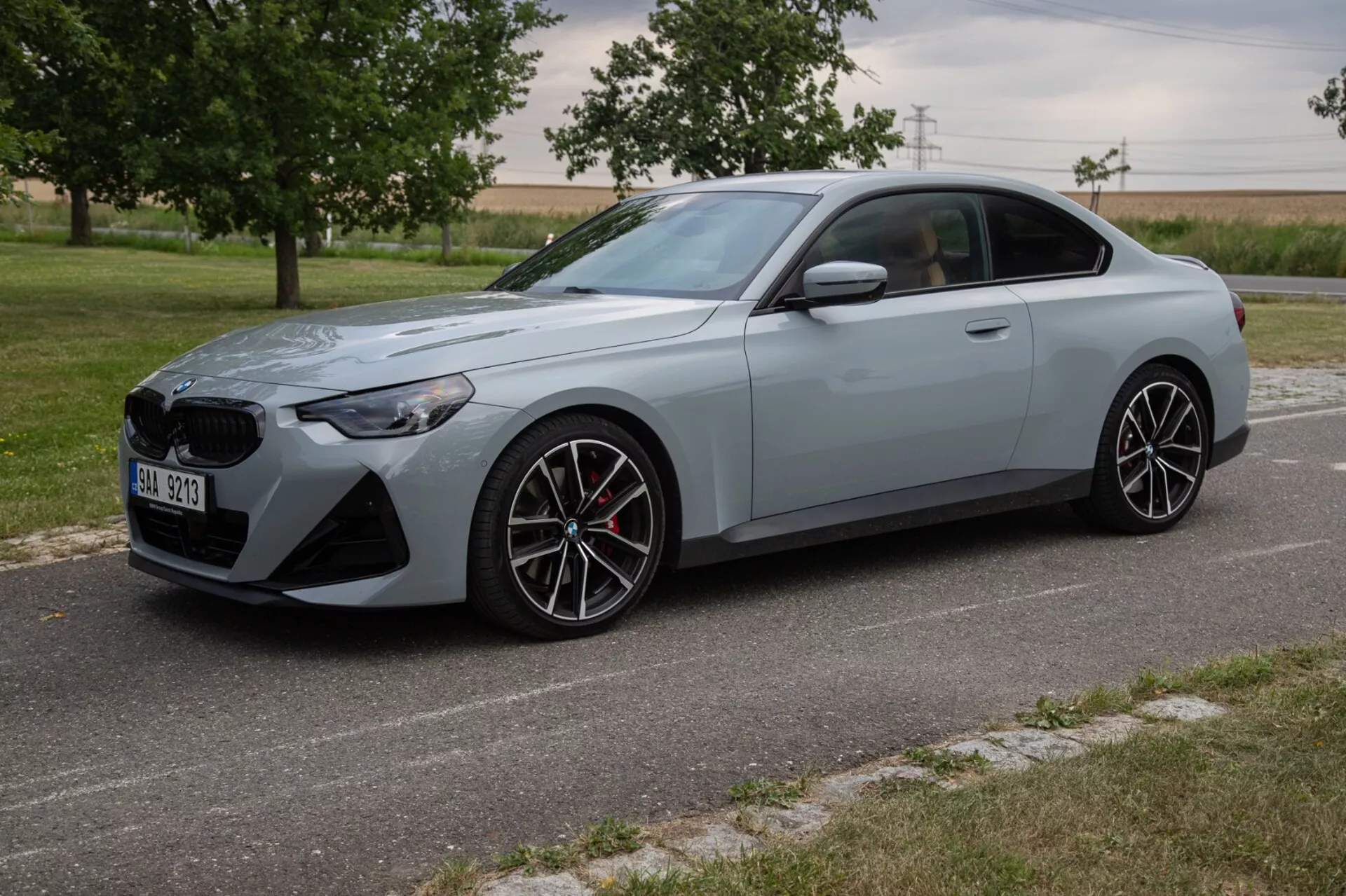 L'image montre un coupé BMW Série 2 de 2022 sous un angle avant trois quarts, mettant en valeur son design extérieur élégant et sportif avec une grande calandre avant et des phares LED angulaires. Le coupé sportif, peint en gris clair mat, est équipé de jantes en alliage à plusieurs rayons, d'un système de freinage haute performance avec des étriers rouges et de lignes sculptées pour un profil aérodynamique. La voiture est garée sur une route pavée avec un paysage naturel de champs en arrière-plan, en plein jour.