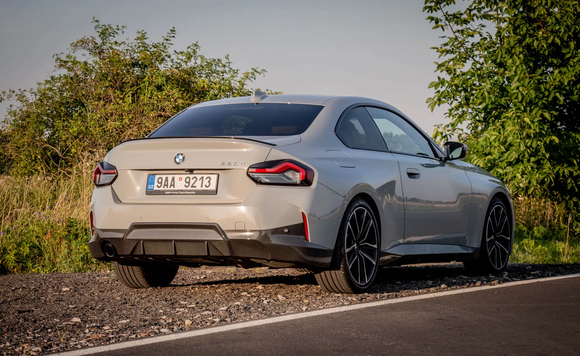 Vue arrière d'une voiture de sport BMW 2er-reihe 2022 (coupé série 2) capturée sous un angle bas. La voiture, garée au bord de la route dans un cadre rural avec des feuillages naturels, est visible sous un éclairage doux. Les insignes distinctifs identifient la marque et le modèle, montrant l'arrière incluant le coffre, le pare-chocs arrière et les feux arrière enveloppants. Les doubles tuyaux d'échappement suggèrent un moteur puissant. Un petit spoiler arrière intégré et de grandes jantes en alliage à rayons fins indiquent un style aérodynamique et performant. La peinture métallisée de la voiture et la ligne de toit doucement inclinée renforcent son aspect haut de gamme. Les caractéristiques modernes visibles possibles incluent les feux arrière à LED, les capteurs de stationnement et une caméra de recul.