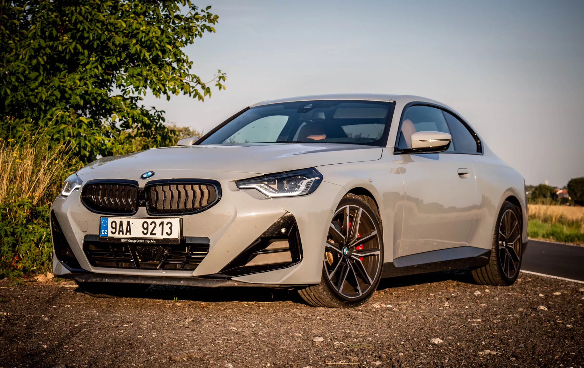 Image d'un coupé sportif moderne BMW Série 2 2022. La voiture est garée sur le côté de la route en plein jour, vue sous un angle oblique depuis l'avant gauche, mettant en valeur le côté conducteur. Les caractéristiques incluent un design aérodynamique élégant, de grandes roues en alliage multibranches avec finition foncée, des étriers de frein contrastants, une calandre distinctive flanquée de phares LED agressifs étroits, une caméra avant au-dessus de la plaque d'immatriculation, de grandes prises d'air dans le pare-chocs avant, une peinture métallique claire, des rétroviseurs latéraux avec clignotants intégrés et une vue partielle du toit panoramique. La voiture est positionnée près de l'appareil photo avec un arrière-plan rural ou suburbain, un éclairage chaleureux et des ombres indiquant un soleil bas le matin ou en fin d'après-midi.