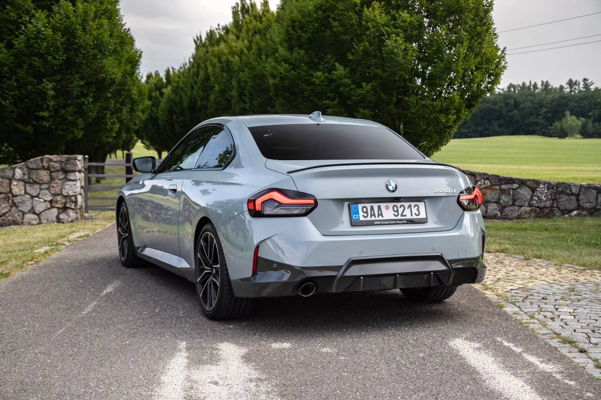 Image de la BMW 2er-Reihe 2022 2-Series Coupé capturée sous un angle de trois quarts arrière. La voiture, positionnée presque en diagonale, présente des feux arrière à LED angulaires distinctifs, deux sorties d'échappement, des roues en alliage à rayons multiples et une finition de peinture métallisée brillante. Le design arrière comporte un emblème du fabricant centré et un badge de version de modèle/moteur sur le couvercle du coffre. L'extérieur comprend une route pavée ou une allée, une zone gazonnée, des arbres et un mur de pierre partiellement visible.
