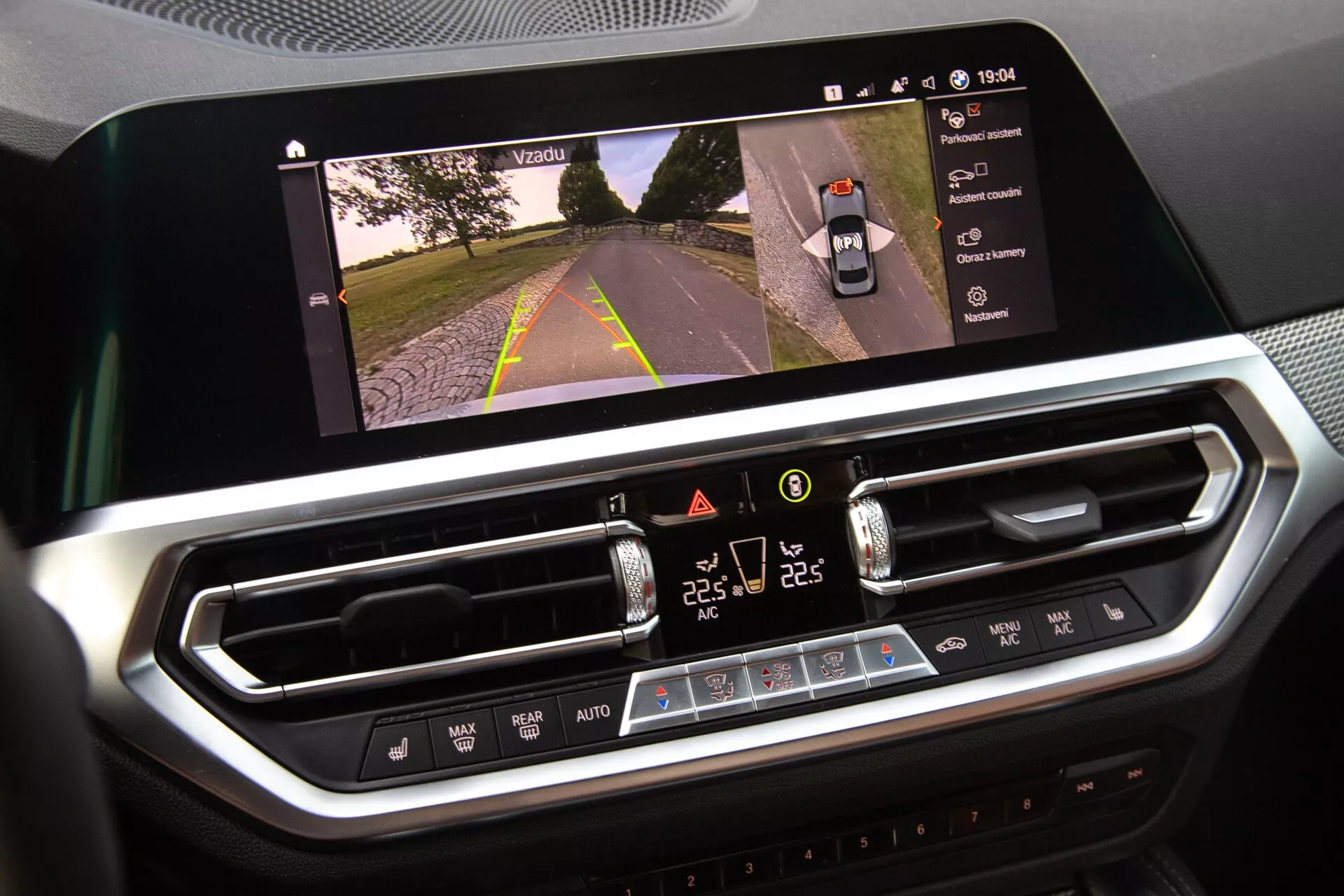 Gros plan de l'intérieur de la BMW 2er-Reihe 2022 montrant l'affichage de l'infodivertissement et le système de contrôle climatique. L'écran tactile horizontal affiche une vue de la route indiquant l'activation de la caméra, tandis que l'interface de contrôle climatique numérique en dessous présente des réglages de température à double zone, de vitesse de ventilateur et des boutons physiques. Un design moderne avec des garnitures chromées suggère des matériaux de haute qualité.