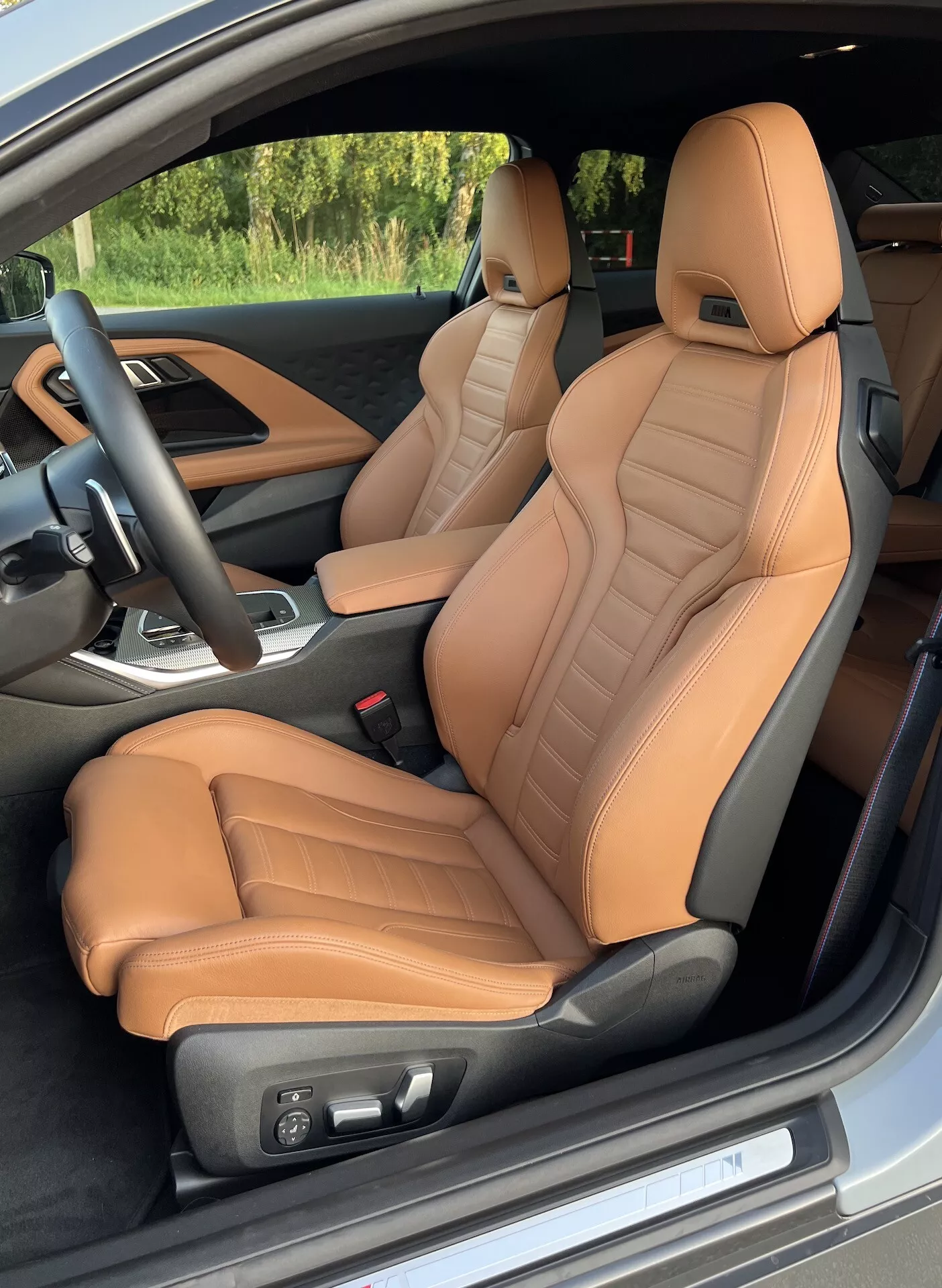 Vue intérieure du BMW Série 2 Coupé 2022 depuis la porte côté conducteur, mettant en évidence les sièges avant rembourrés de cuir coloré bronze avec des détails matelassés et un soutien ergonomique. Les réglages électriques des sièges, le volant noir avec commandes intégrées et une vue partielle de la console centrale avec levier de vitesses suggèrent des caractéristiques de luxe et de performance.