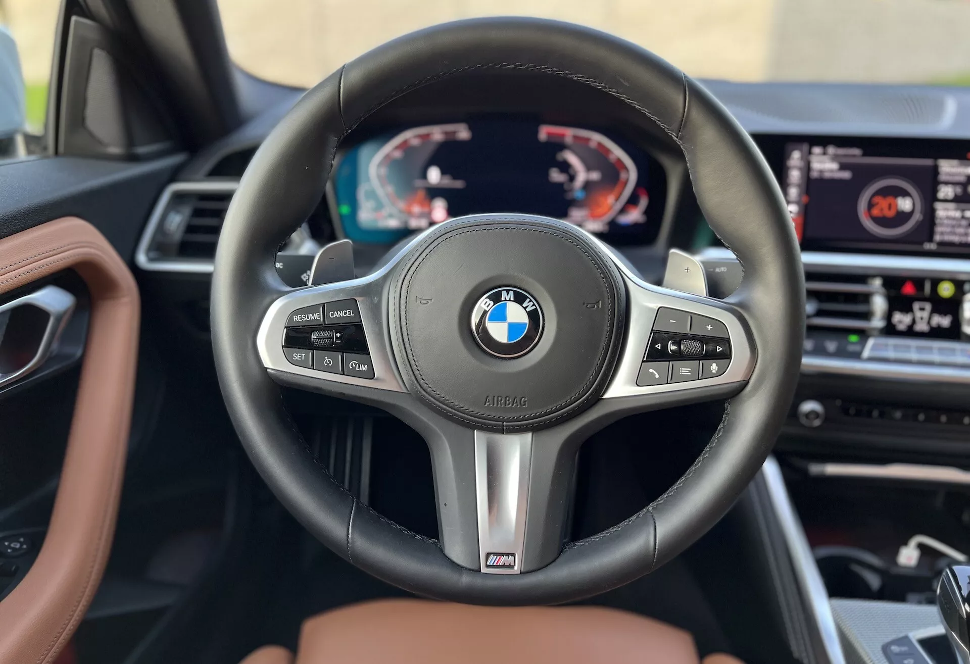 Vue intérieure d'une BMW Série 2 Coupe 2022 se concentrant sur le volant et le tableau de bord depuis la perspective du siège conducteur. Comporte un volant à trois branches BMW avec des boutons multifonctions pour le régulateur de vitesse, la gestion audio et la téléphonie, ainsi que des palettes de changement de vitesse et un emblème de performance 'M'. Derrière, un tableau de bord numérique affiche un compte-tours, un compteur de vitesse et des informations multifonctions. Le système d'infodivertissement est visible au centre du tableau de bord, et la finition intérieure comprend du cuir et du bois, suggérant un modèle premium.