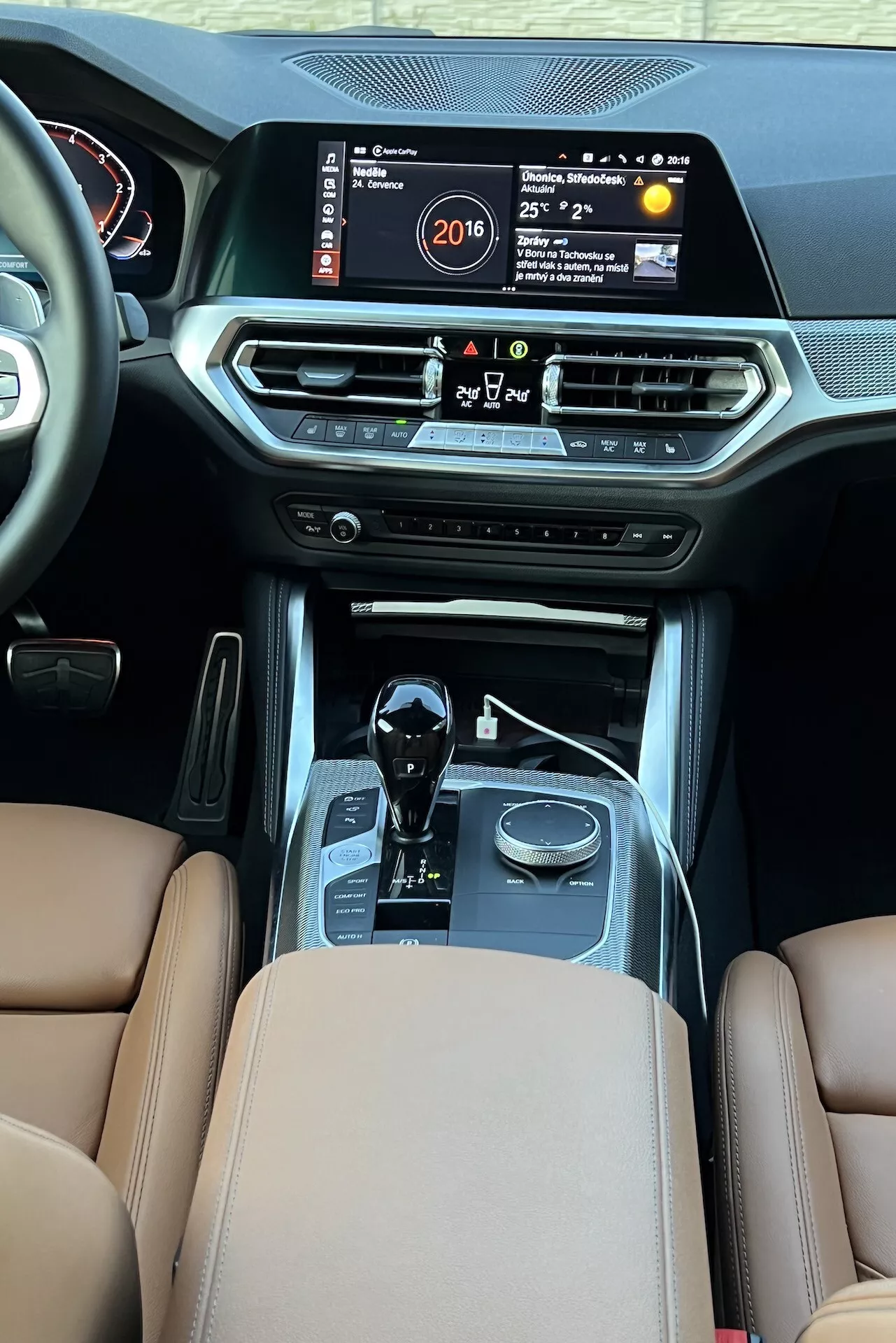 Vue intérieure de la BMW Série 2 2022 depuis la zone arrière des passagers, mettant en évidence la console centrale avec un grand écran d'infodivertissement affichant l'heure, la température et le système multimédia. Les caractéristiques comprennent des bouches de ventilation de la climatisation, des boutons de contrôle du climat, un bouton start/stop, un levier de vitesse automatique avec des accents chromés, des sélecteurs de mode de conduite, des sièges en cuir marron avec une sellerie premium et des coutures, indiquant le luxe et la technologie automobile avancée.