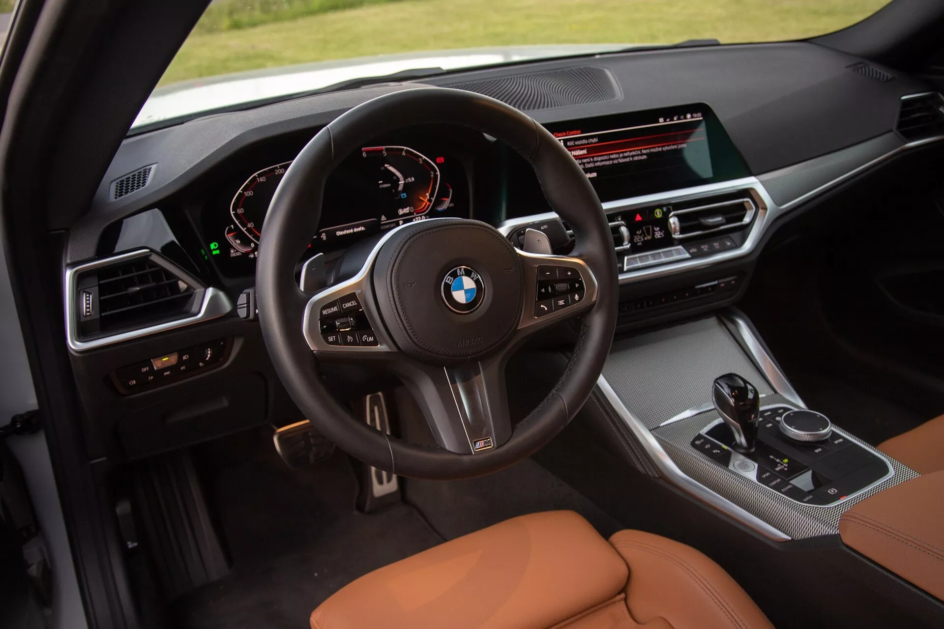 Image intérieure d'une BMW Série 2 Coupé 2022 se concentrant sur le cockpit du conducteur. La prise de vue élevée offre une vue détaillée du volant multifonction avec logo BMW, du tableau de bord numérique affichant le compteur de vitesse et le compte-tours, et de la console centrale orientée conducteur avec un large écran d'infodivertissement, des boutons de contrôle climatique, le levier de vitesses et la molette de commande iDrive. Les sièges sont en cuir couleur caramel ou brun clair, mettant en avant le luxe et le confort.