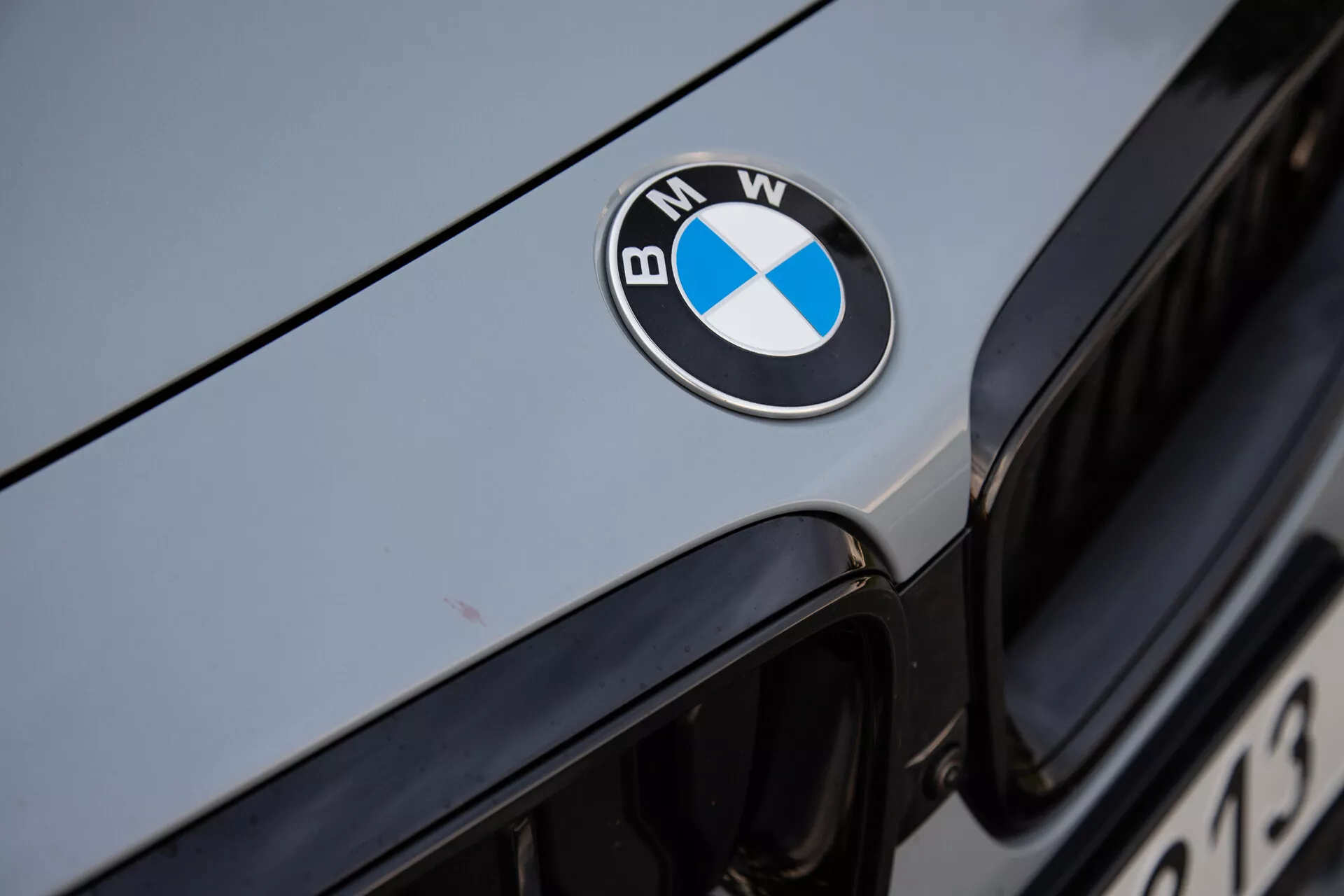 Image en gros plan de l'avant de la BMW Série 2 Coupé 2022, avec un badge BMW centré en noir, bleu et blanc sur le capot. L'image capture le bord supérieur de la grille de calandre iconique en arrière-plan, avec un extérieur de voiture métallique de couleur argentée ou gris clair reflétant la lumière ambiante. La mise au point nette sur le badge met en valeur la marque de luxe et la surface bien entretenue du véhicule.
