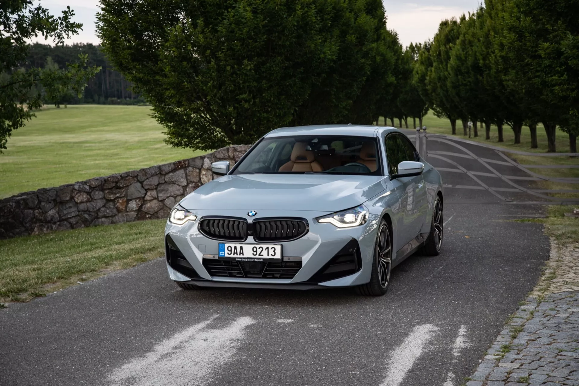 Image détaillée d'une BMW Série 2 Coupé 2022 capturée sous un angle trois-quarts avant, montrant ses profils avant et latéral sur une route en asphalte tranquille bordée d'arbres taillés et d'un mur de pierre. La calandre emblématique en forme de rein de BMW et les élégants phares à LED ornent la face avant, tandis que les prises d'air sur le pare-chocs renforcent son look sportif. Les lignes profilées du capot suggèrent la performance, les ailes bien définies indiquent la puissance et les jantes en alliage à plusieurs branches dénotent une finition sportive. Le profil latéral présente une ligne caractéristique qui donne une impression de mouvement, des rétroviseurs avec clignotants intégrés et une lueur de toit ouvrant. L'angle standard du pare-brise et les vitres latérales claires ajoutent à l'aspect équilibré. L'éclairage doux, probablement au petit matin ou en fin d'après-midi, crée des ombres subtiles et une présentation soignée du design et de la couleur de la voiture.